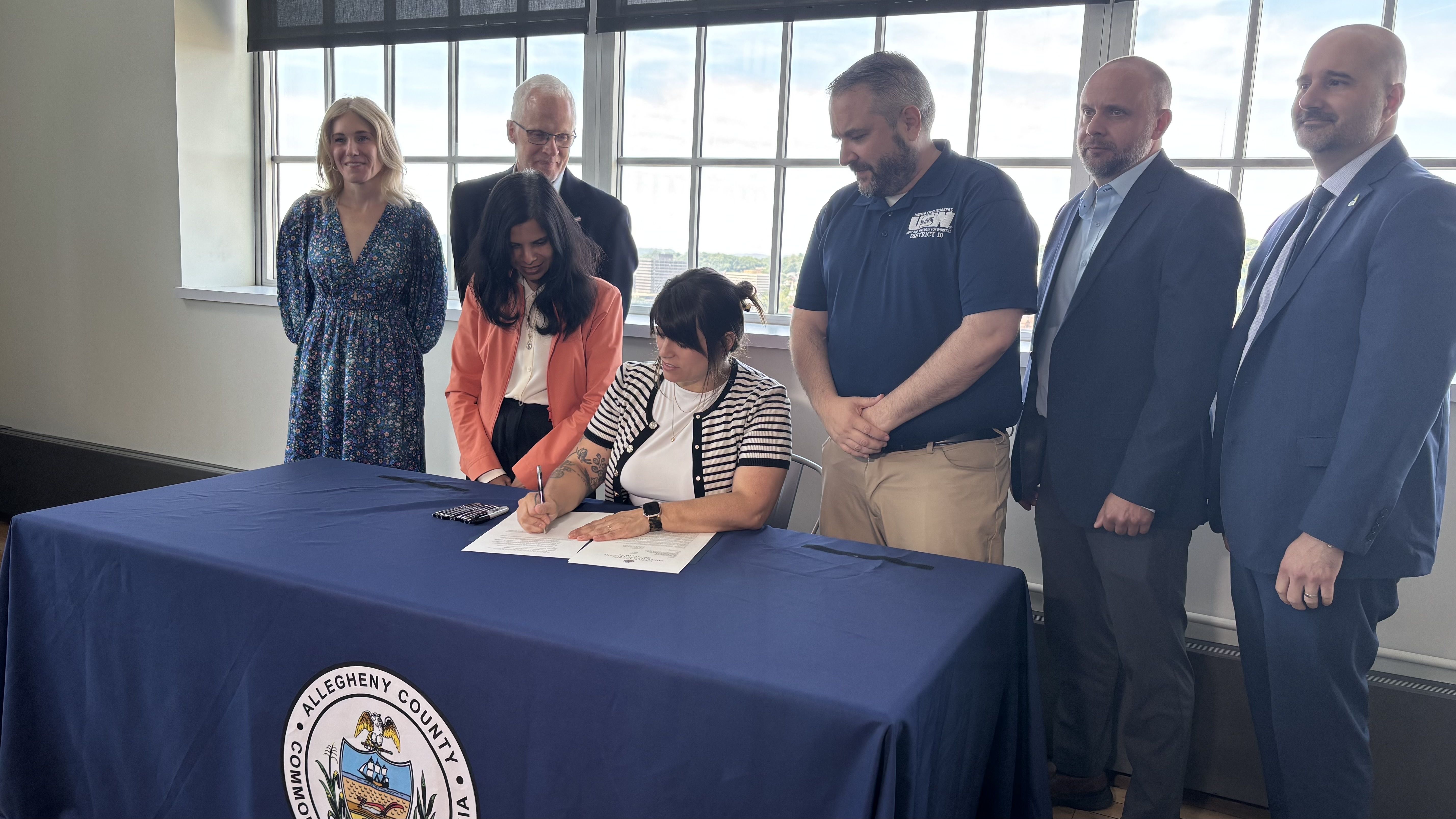 A woman seated at a blue-clothed table signs documents while six people in business attire look on; windowed office setting and the Allegheny County seal on the table are visible.
