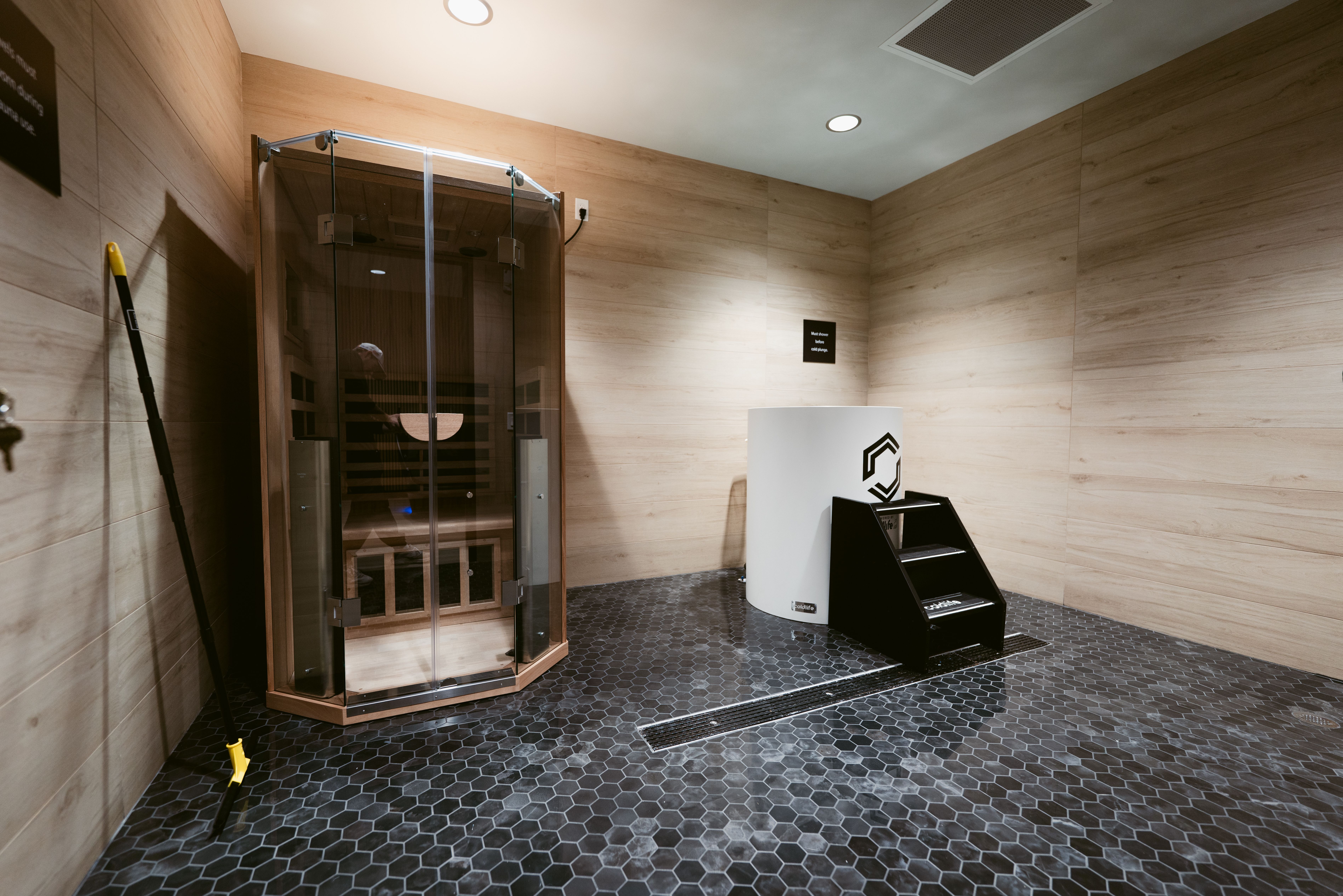Spa sauna room with wood-paneled walls, a glass-front sauna on the left, dark hex tile floor, and a white curved reception stand with a black logo on the right; a yellow-handled cleaning tool leans against the wall.