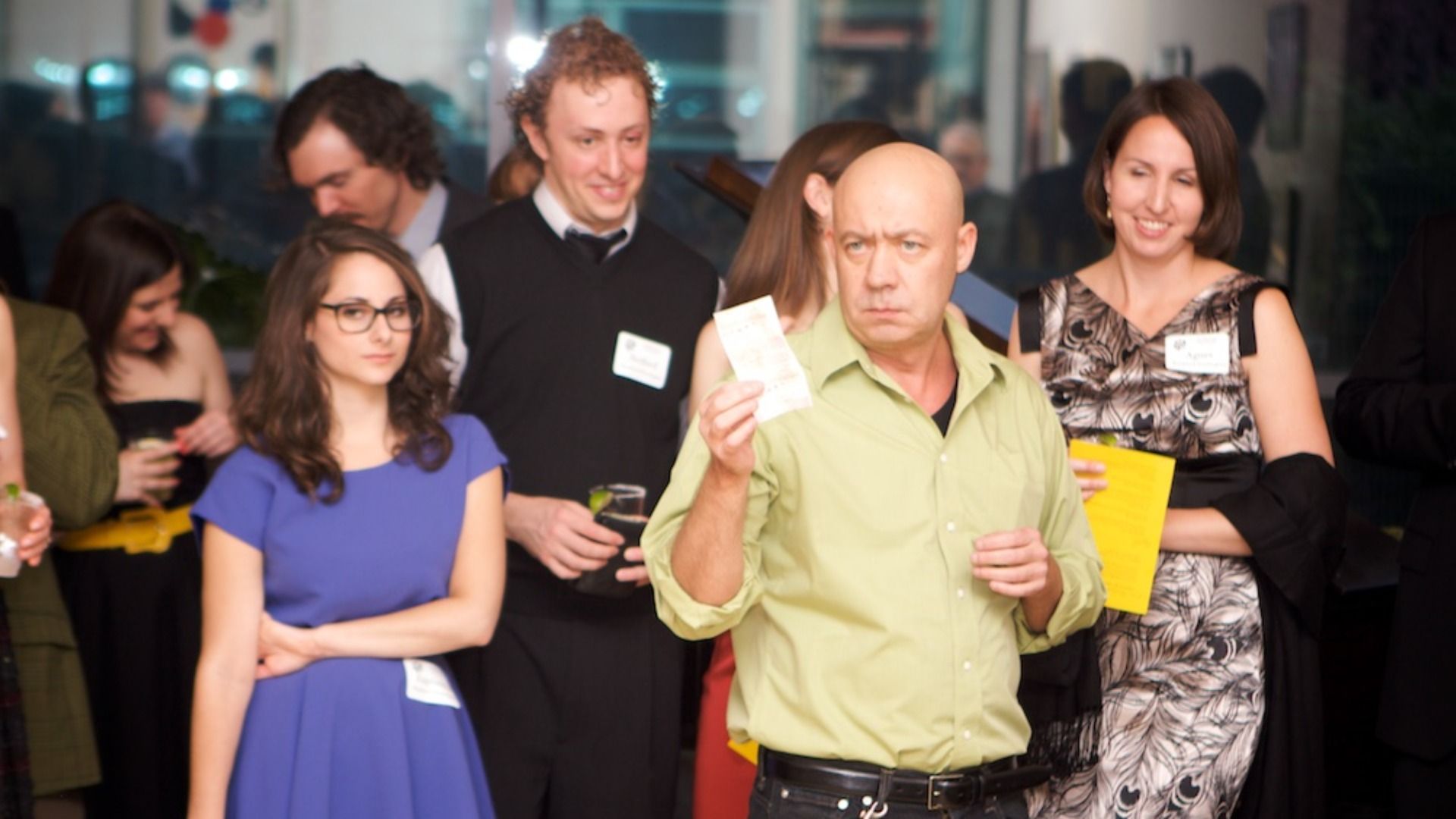 Group of adults at an indoor event. Foreground: a bald man in a pale green shirt holds a ticket; beside him, a woman in a blue dress with glasses. Others smile and mingle in the background.