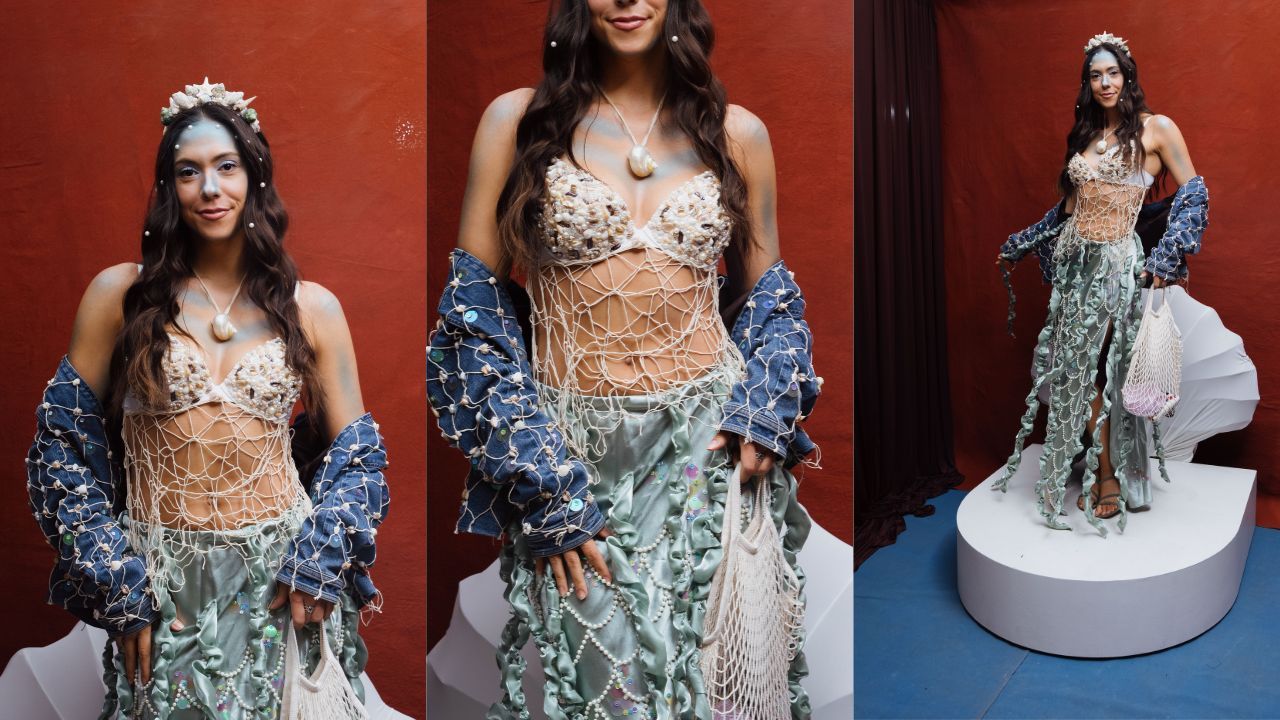 Model stands on a white pedestal against a red wall, wearing a seashell bikini top with a net overlay and pale green layered skirt, pearl jewelry, a shell necklace, a flower crown, and a net bag.