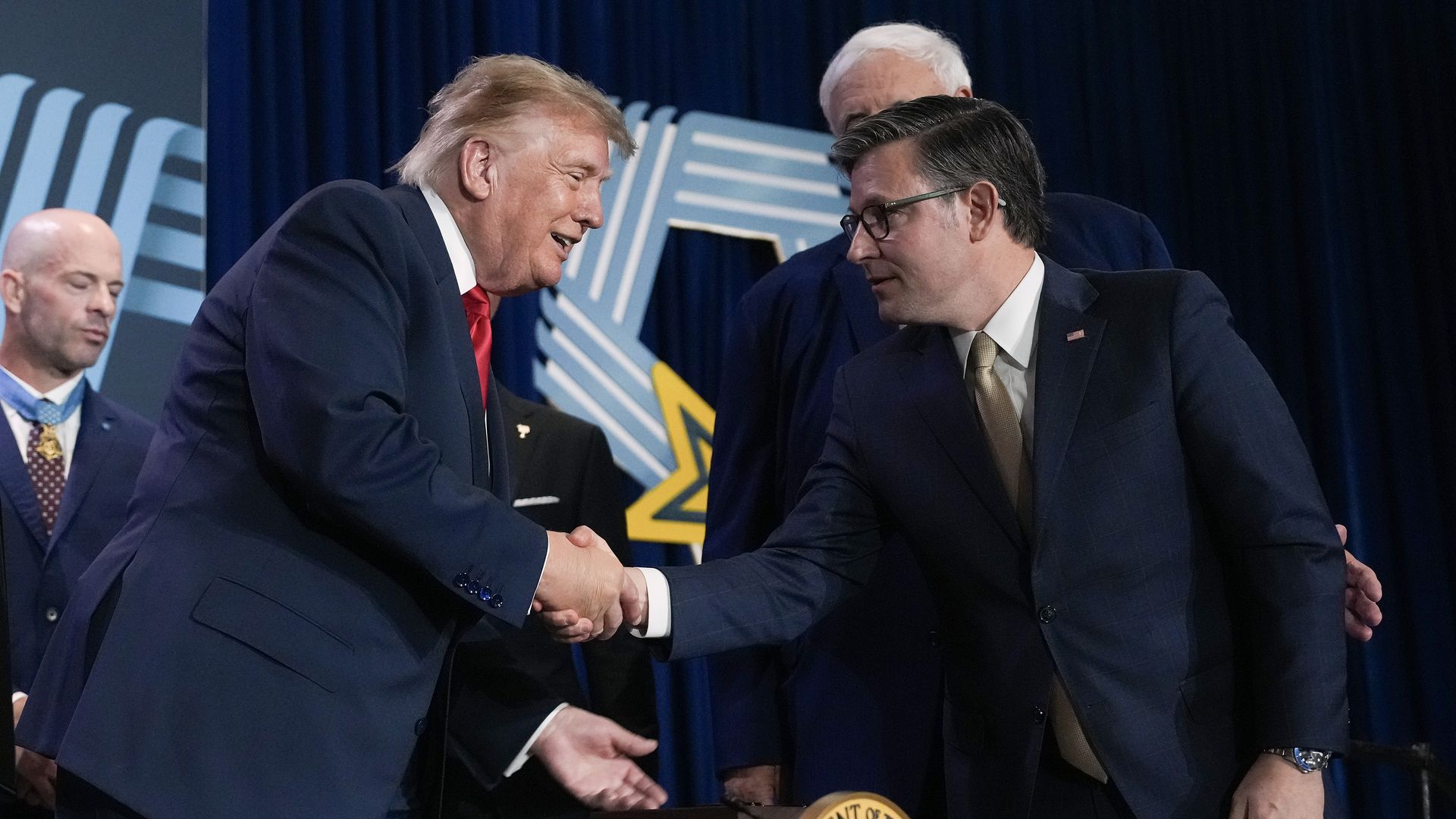Trump, wearing a dark suit, a white collared shirt and a red tie, leans across a desk as he shakes hands with Speaker Johnson, wearing a dark suit, a white collared shirt and a gold tie.