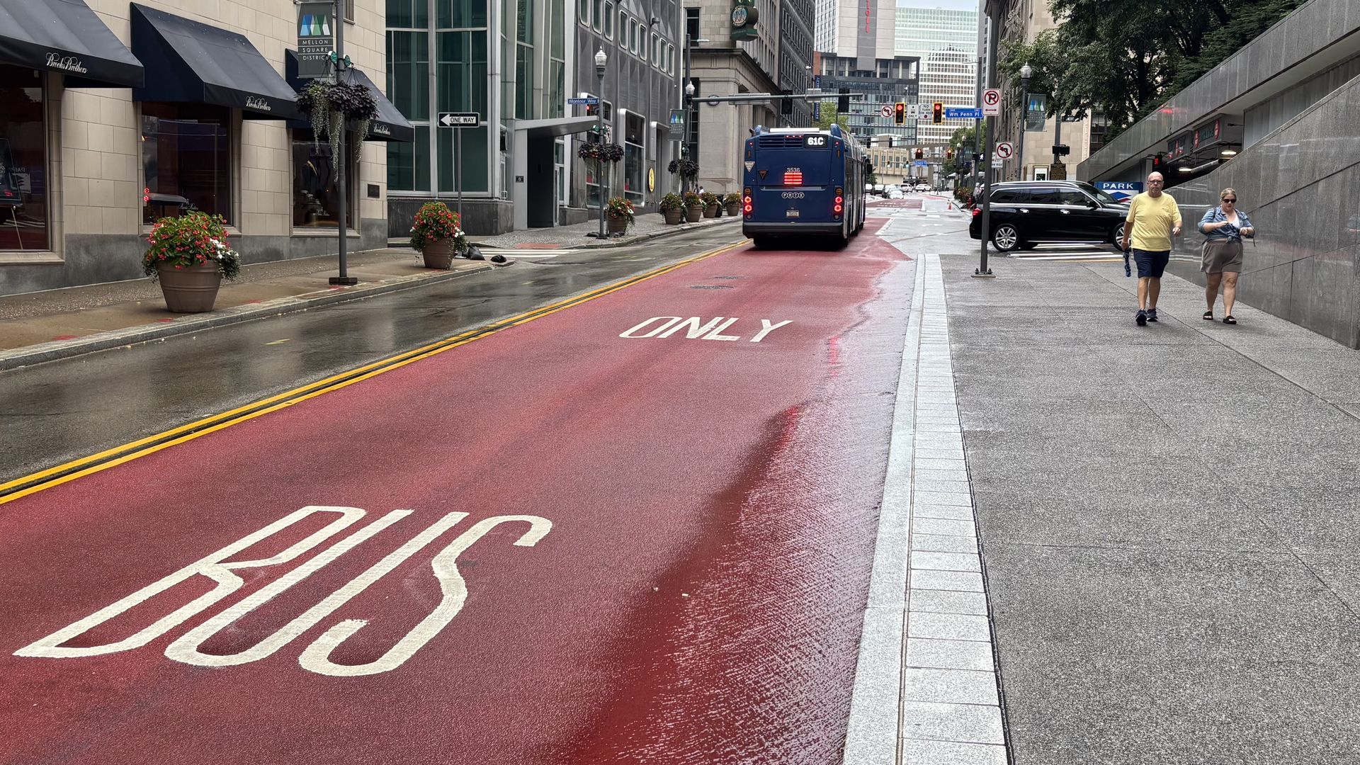 Wet city street in Pittsburgh with a red bus-only lane, a blue bus labeled 61C, two pedestrians walking on the gray sidewalk, and beige buildings with black awnings and flower pots.