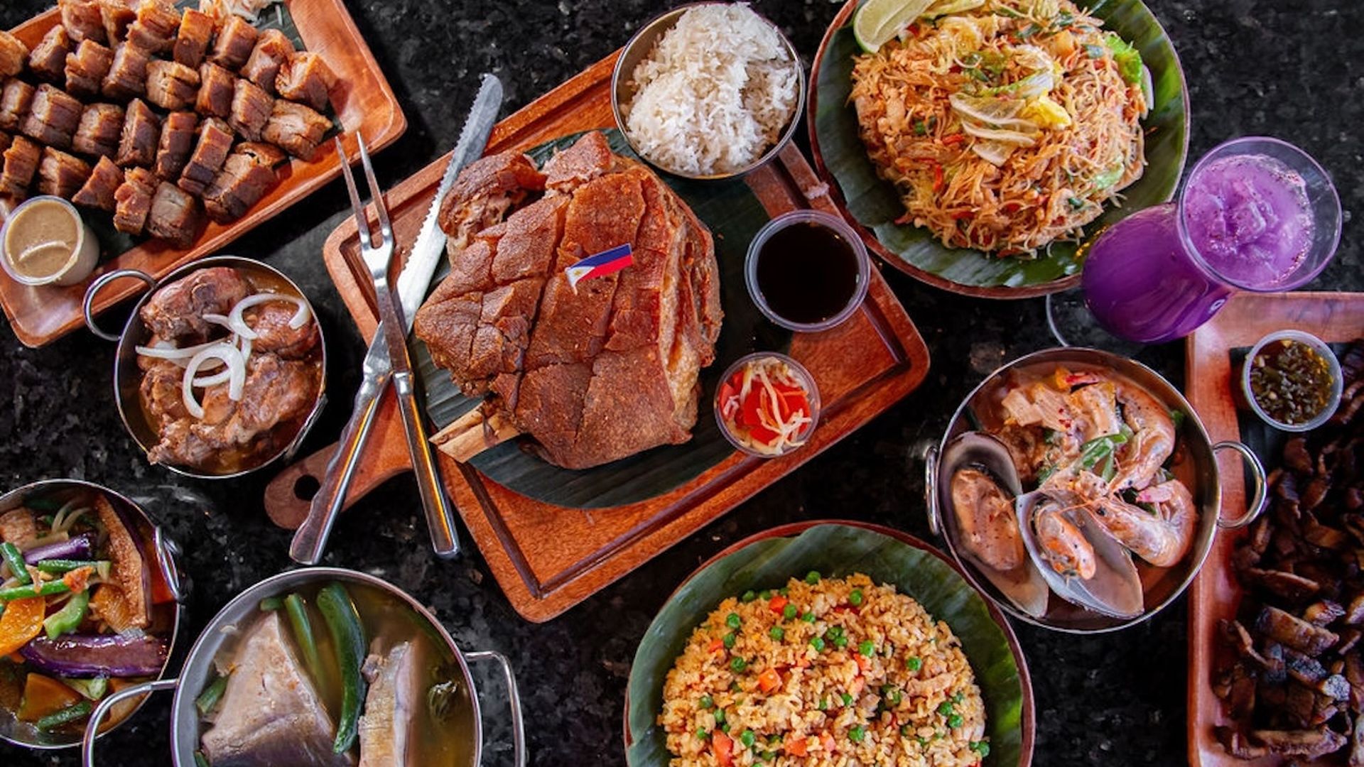 Top-down feast: center roast lechon on cutting board, surrounded by fried rice, pancit, clams in broth, beef stew, roasted pork cubes, mixed vegetables, a purple drink, and sauces on a dark counter.