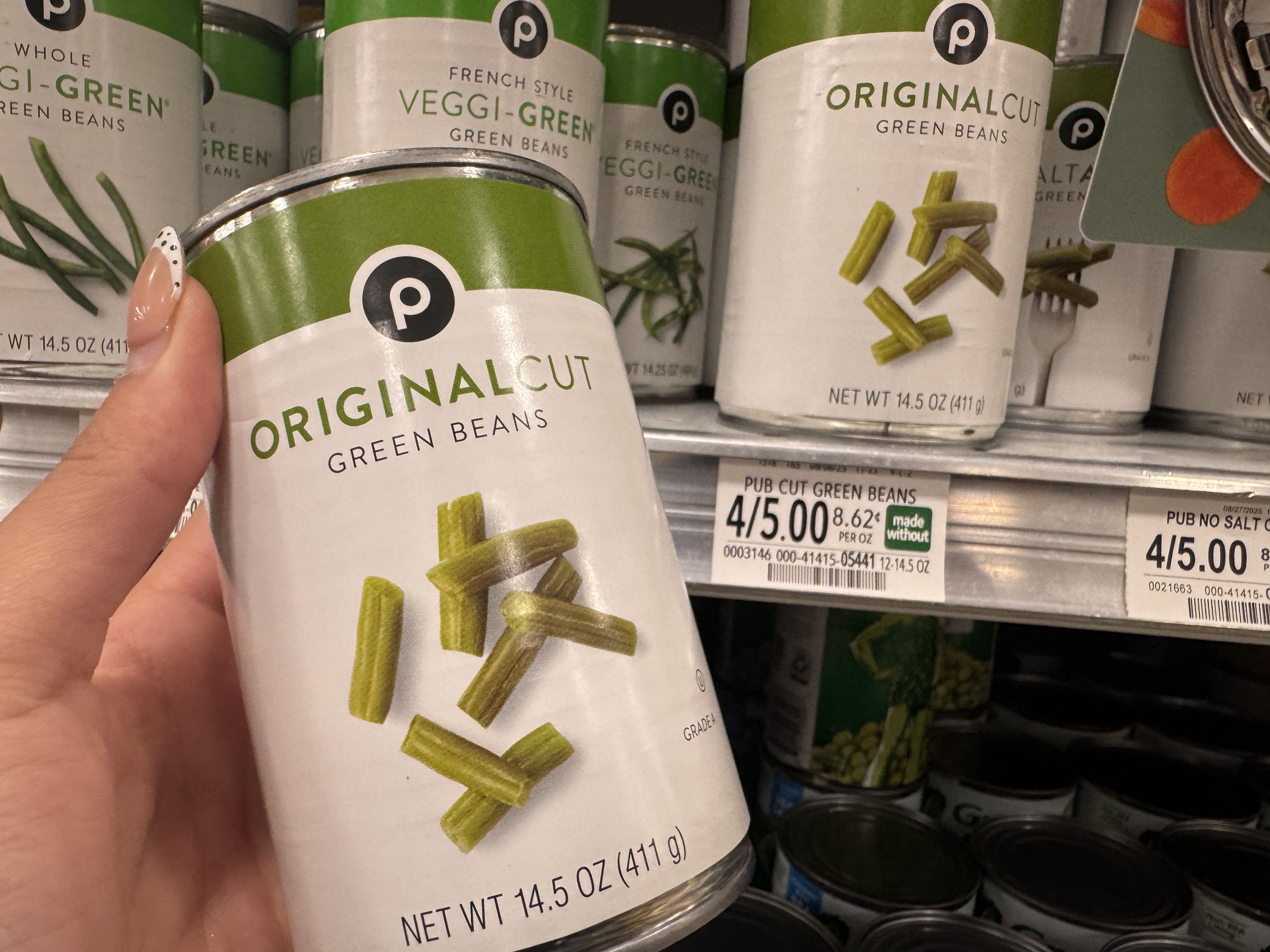 Hand holding a can of Publix Original Cut green beans, 14.5 oz, on a grocery store shelf with similar cans and a price tag of 4 for $5.00.