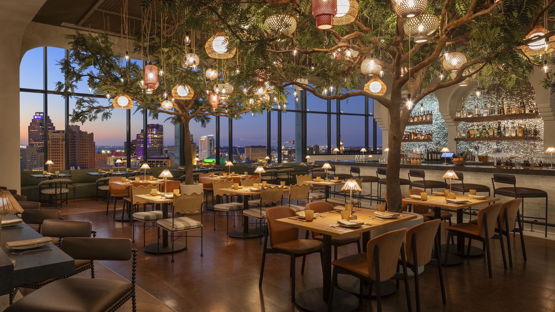 Stylish rooftop restaurant at dusk with large windows showing city skyline, trees with hanging lanterns, warm lighting, wooden tables, chairs, and a well-stocked bar.