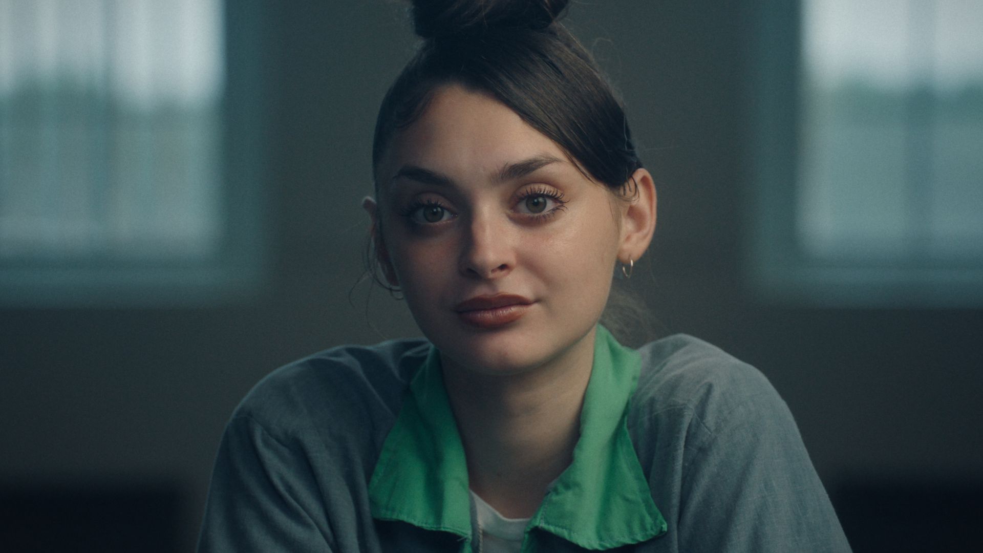 Portrait of a young woman with dark hair in a bun, wearing a green collared shirt over a gray top, looking softly at the camera in a dim room with blurred windows behind her.