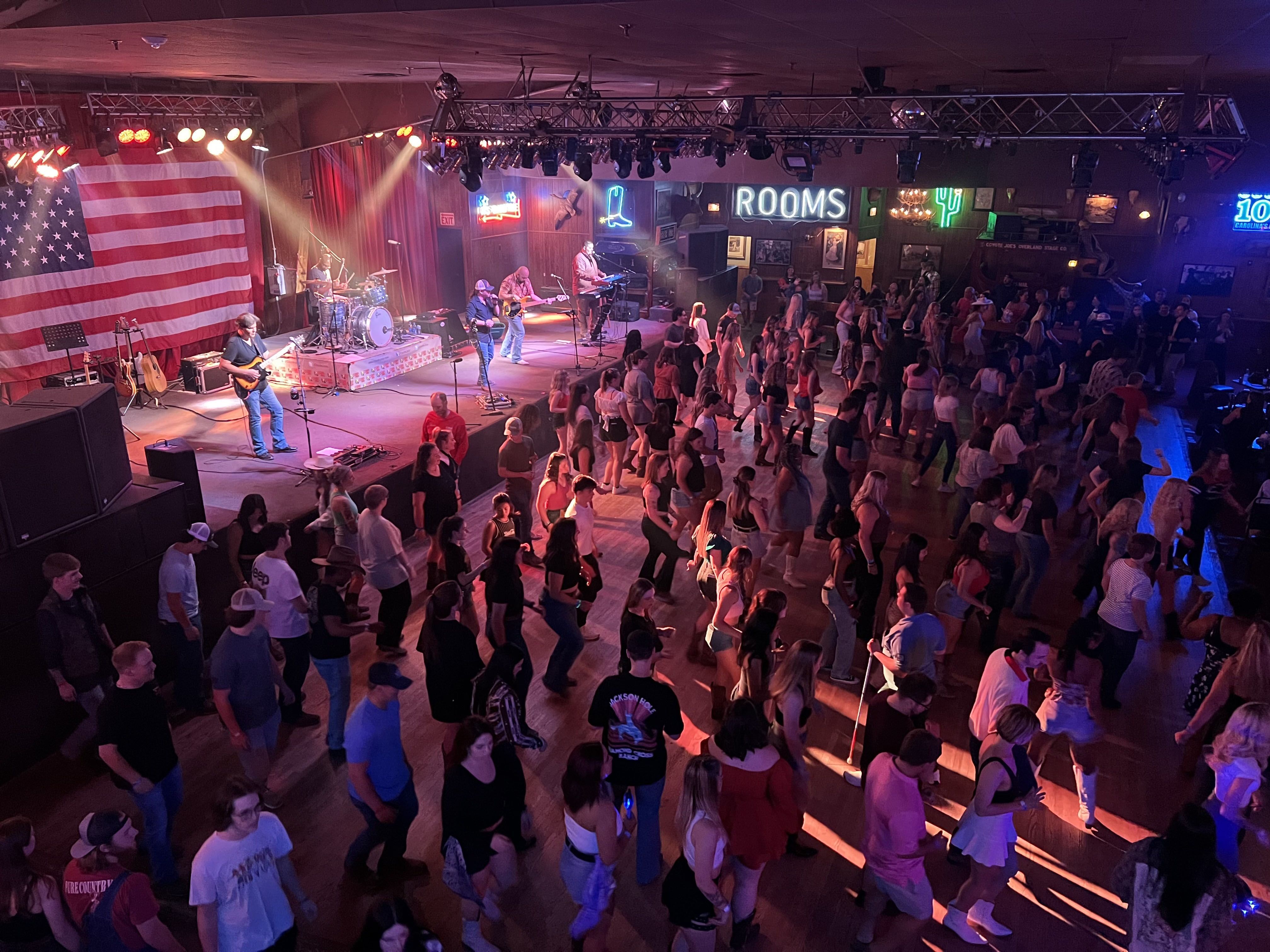Crowded indoor dance floor with people line dancing as a band plays on stage under red and white lights, large American flag backdrop, neon signs reading "ROOMS" and a cactus.
