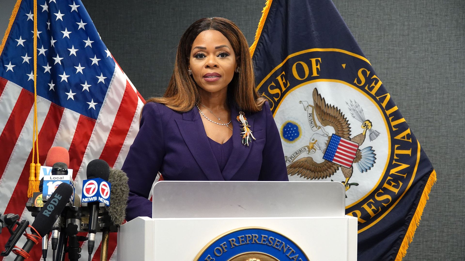Rep. Sheila Cherfilus-McCormick, wearing a purple suit and standing in front of two flags and a gray wall behind a white podium and a cluster of microphones.