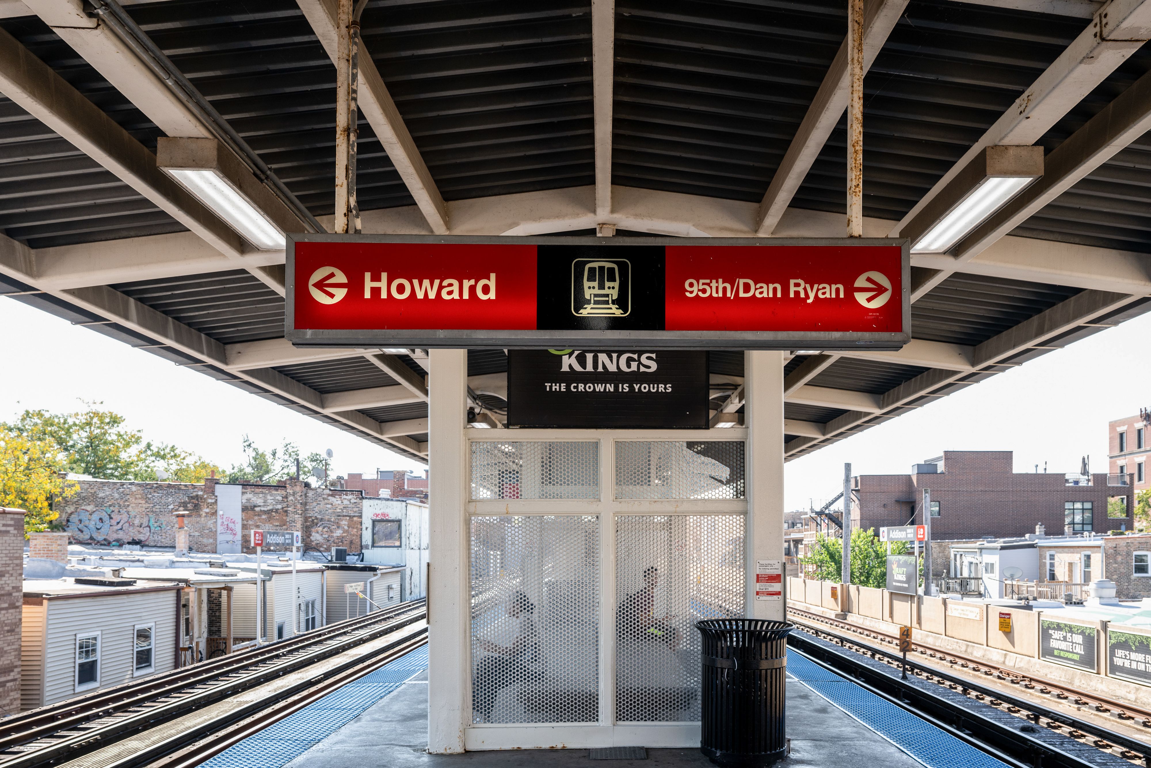 Chicago elevated train platform with sign pointing left to Howard and right to 95th/Dan Ryan stations, under a metal roof with urban buildings in background.