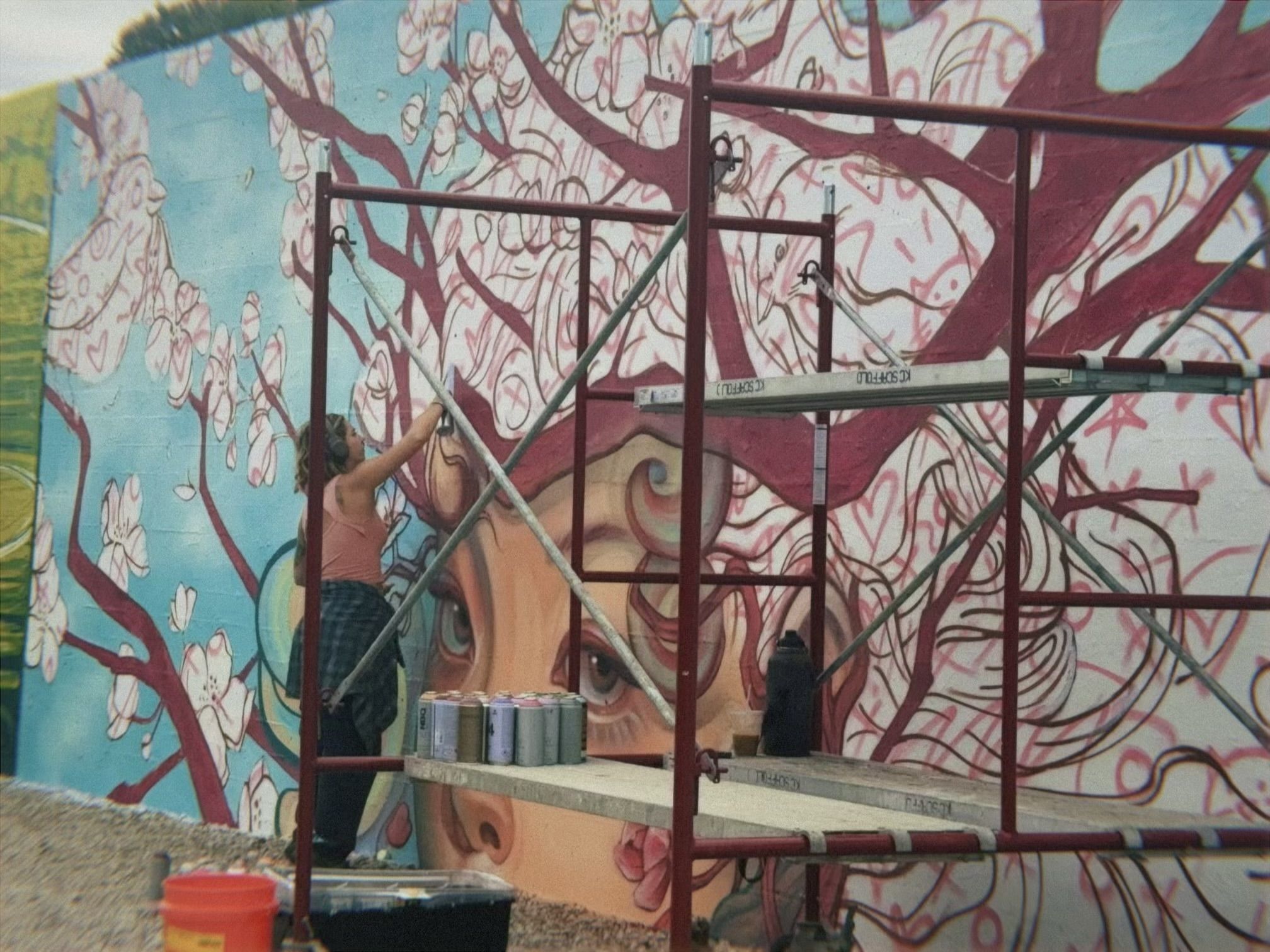 An artist wearing a pink top and headphones painting a large mural of a woman's face with flowing hair that turns into branches and white flowers on a blue background.