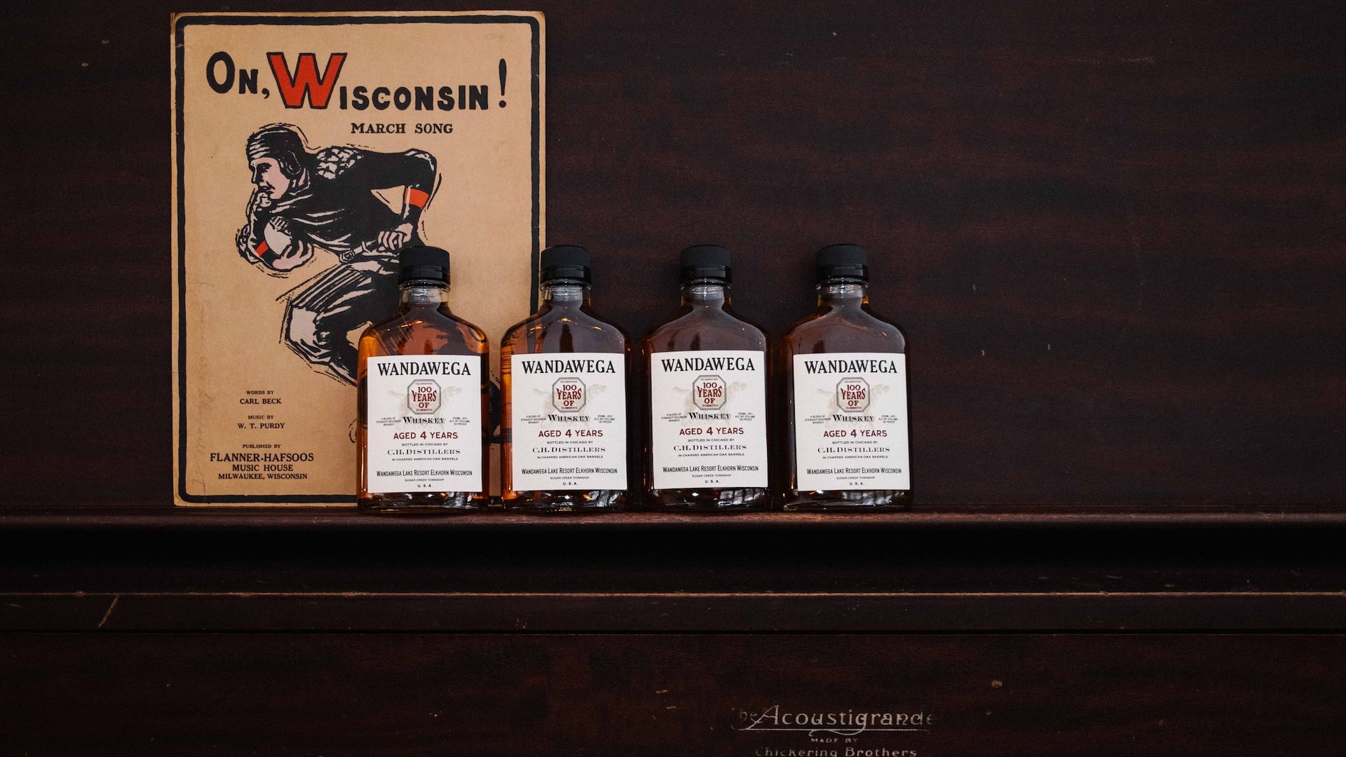 Four bottles of bourbon sitting on a piano in front of a vintage sign with a football player that reads "Oh, Wisconsin!"