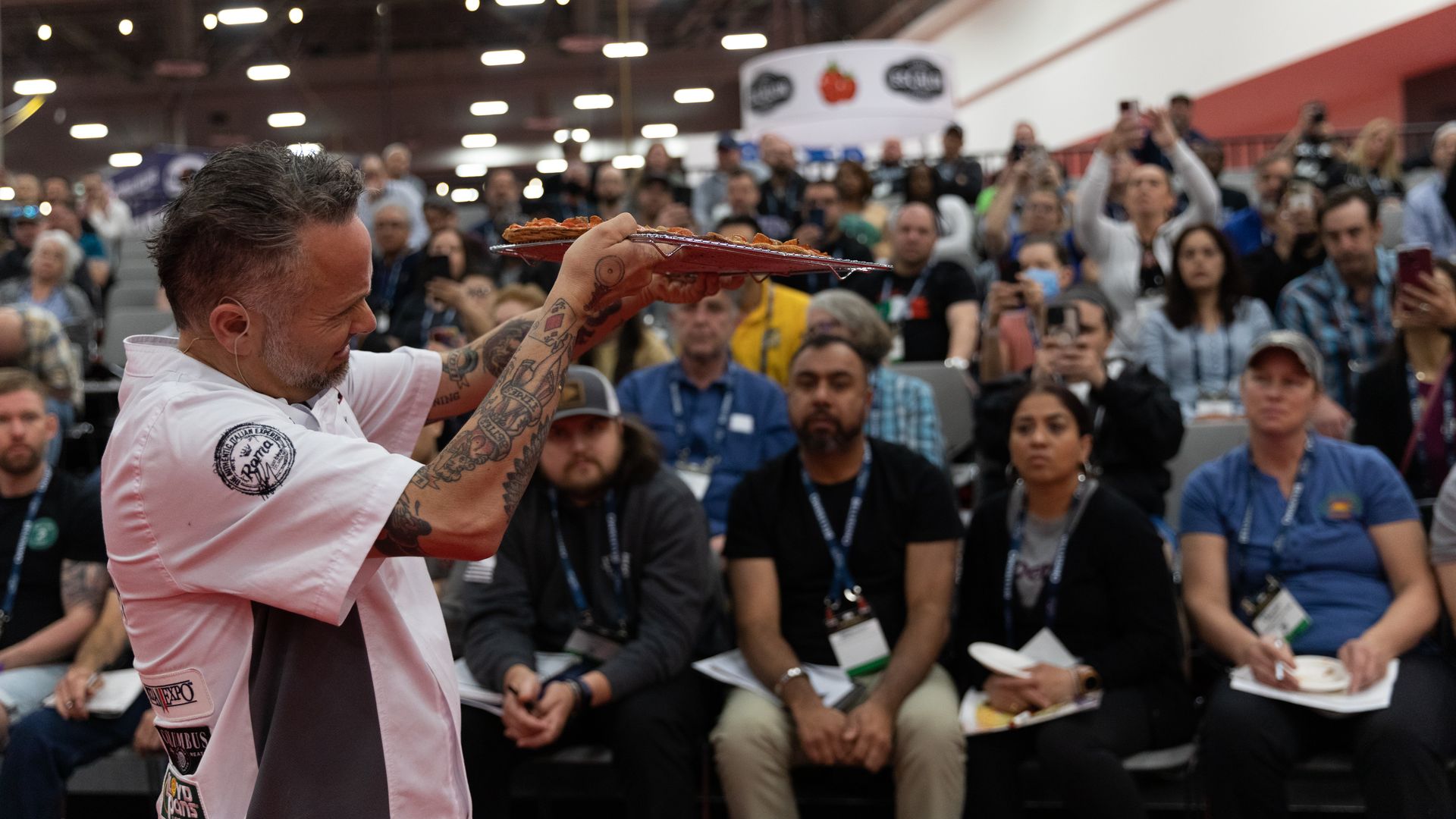 Tattooed chef in a white shirt presents a pizza on a metal tray to a seated audience at an indoor event, with many people focused and some taking photos.