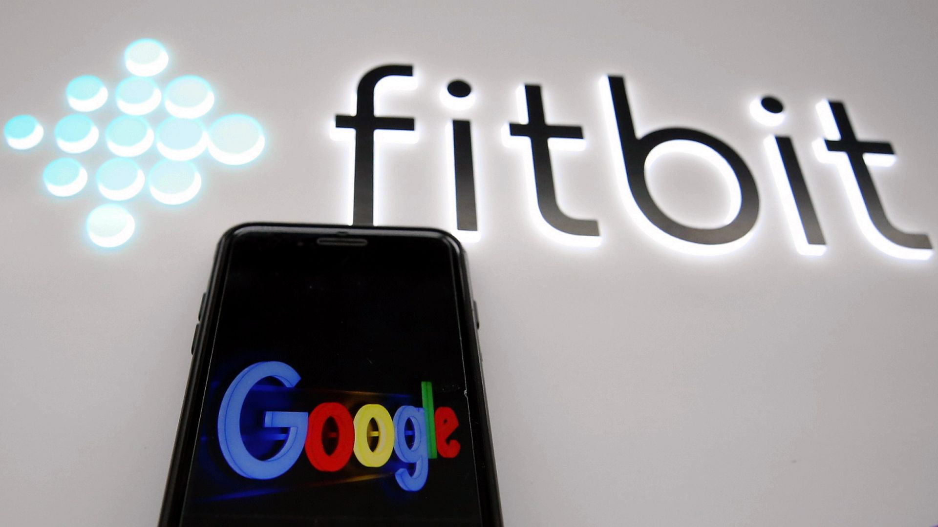 A photo of a smartphone displaying the Google logo, with an illuminated Fitbit sign behind it