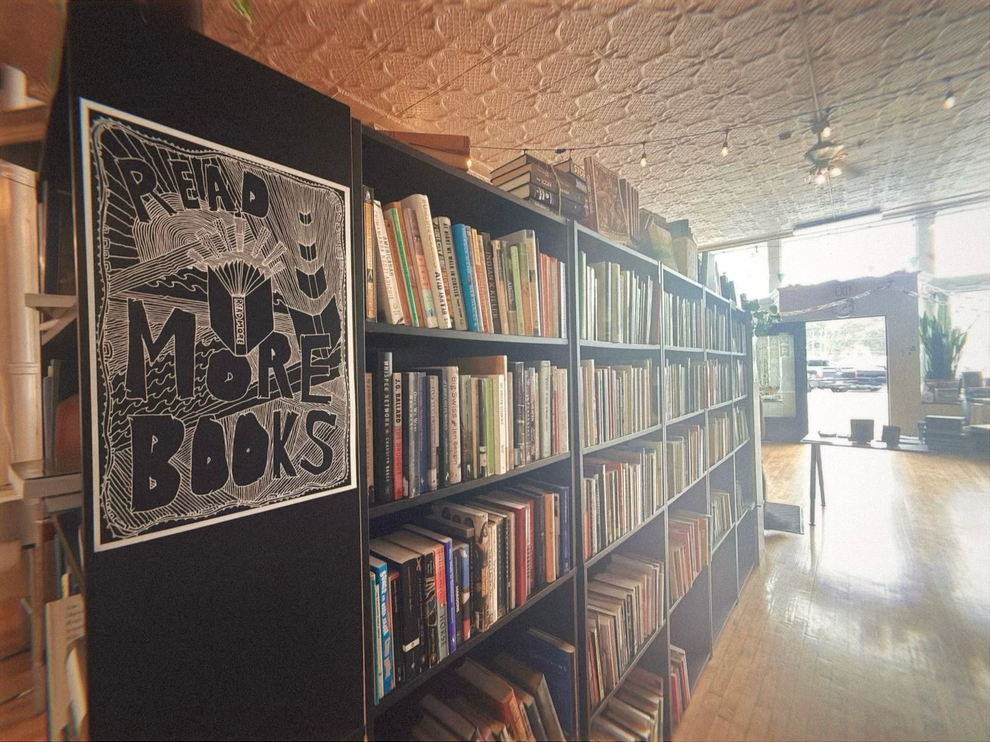 Interior of a bookstore or library with black bookshelves filled with books. A black-and-white poster on the end shelf reads "READ MORE BOOKS." Light wooden floor and patterned ceiling visible.