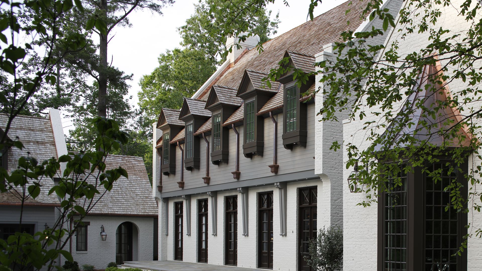 White brick large house with brown window frames, a wooden shingle roof, dormer windows, surrounded by greenery and trees with a curved concrete driveway.