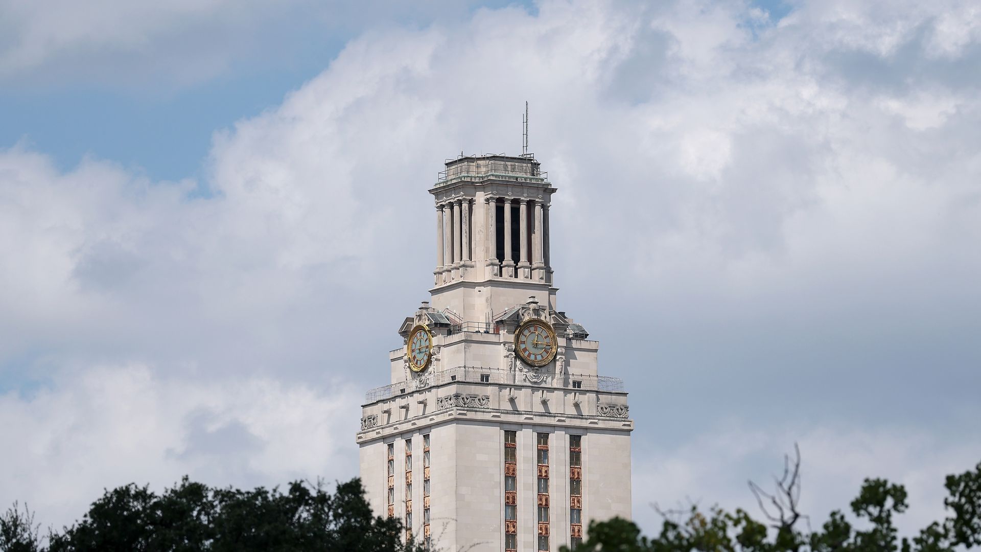 Tall beige clock tower with large round clocks on each side, partly obscured by trees, under a cloudy blue sky.