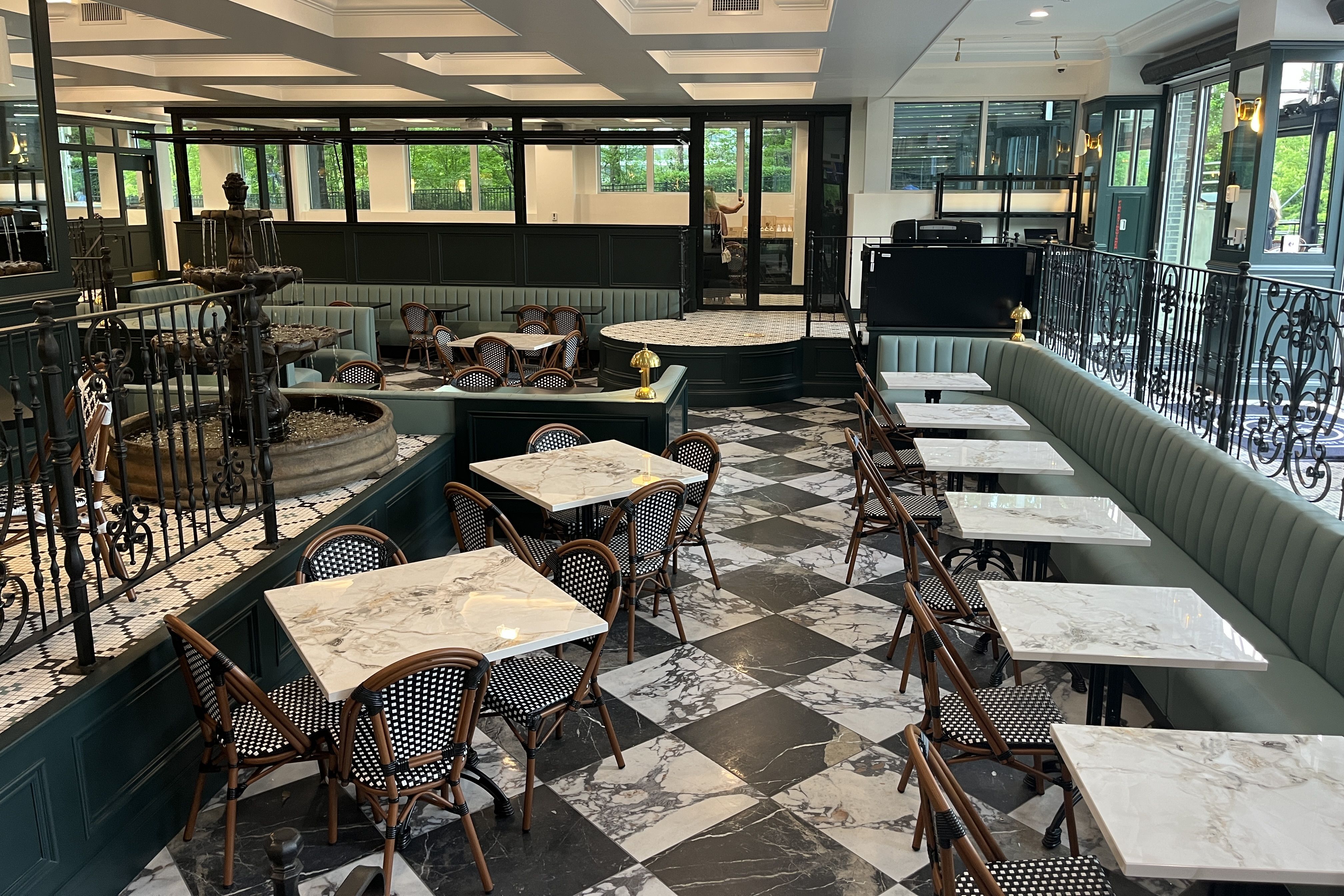 Elegant cafe interior with marble-top tables, wicker chairs, teal banquettes, black paneled walls, ornate wrought-iron railing, a central fountain, and large windows showing greenery outside.