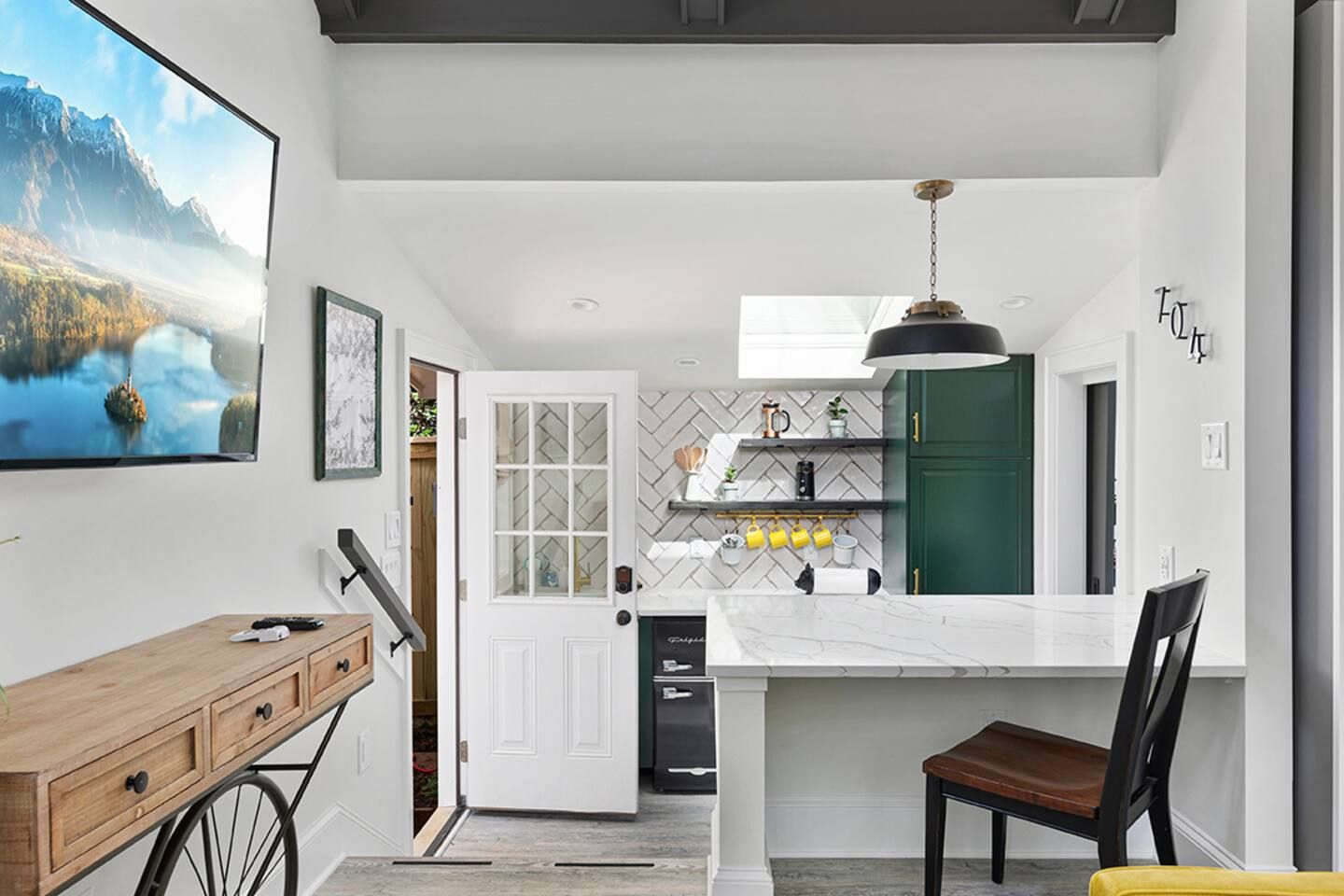 Modern kitchen with green cabinets, white marble countertop island with black chair. Yellow mugs hanging, white herringbone tile backsplash, skylight, black pendant light, wooden console table.