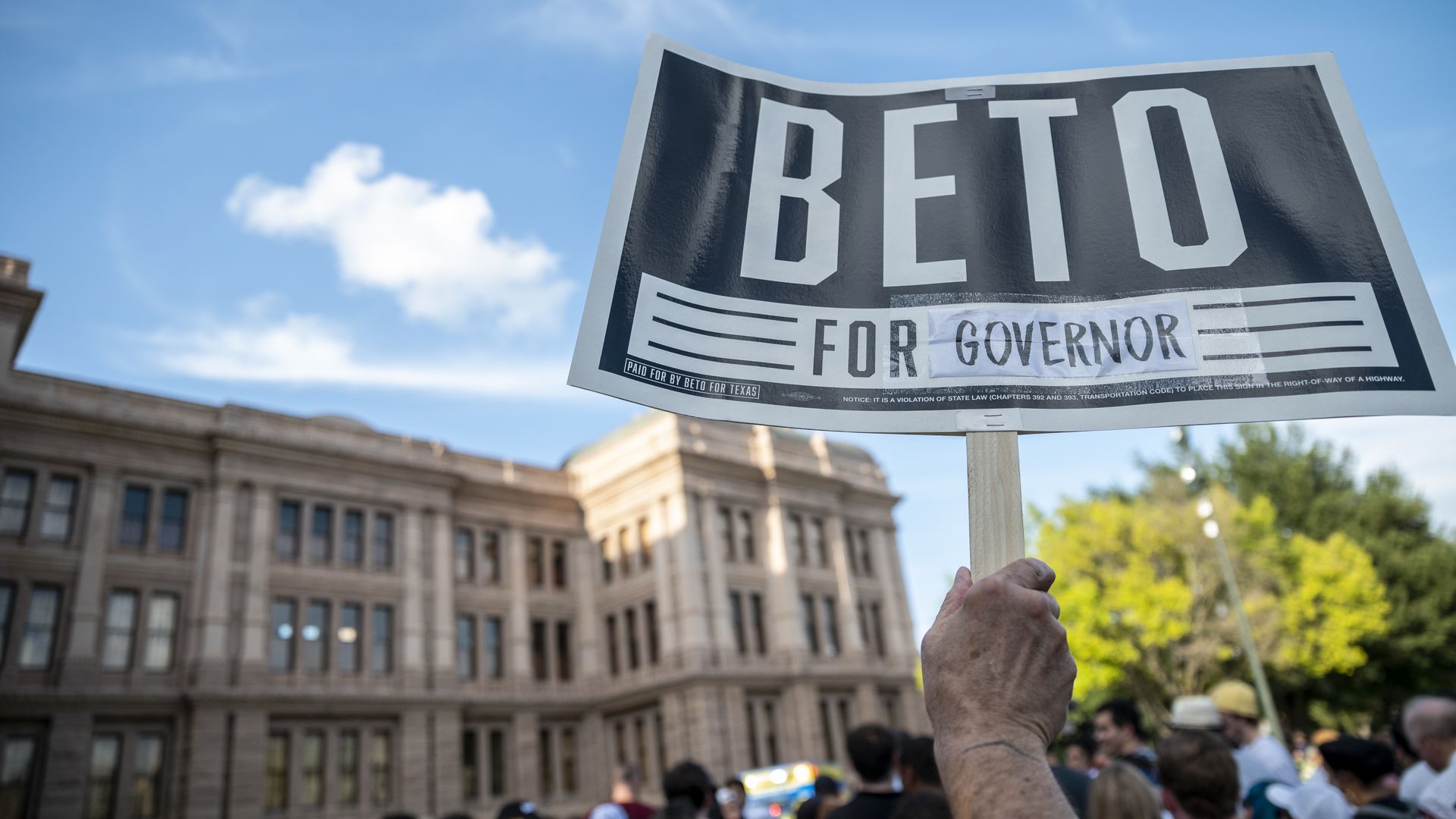 A photo of a hand holding a sign that reads "Beto For Governor" in the foreground of the Texas Capitol