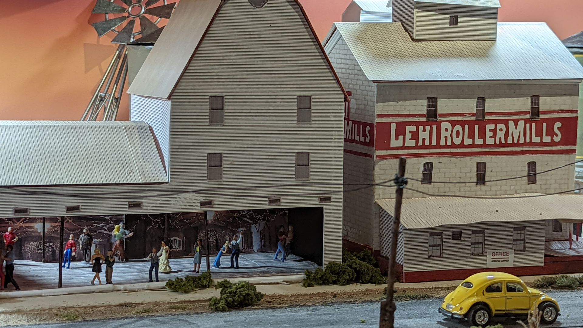 Miniature figurines of people dance in a barn labeled "Lehi Roller Mills" with a miniature yellow VW bug outside.