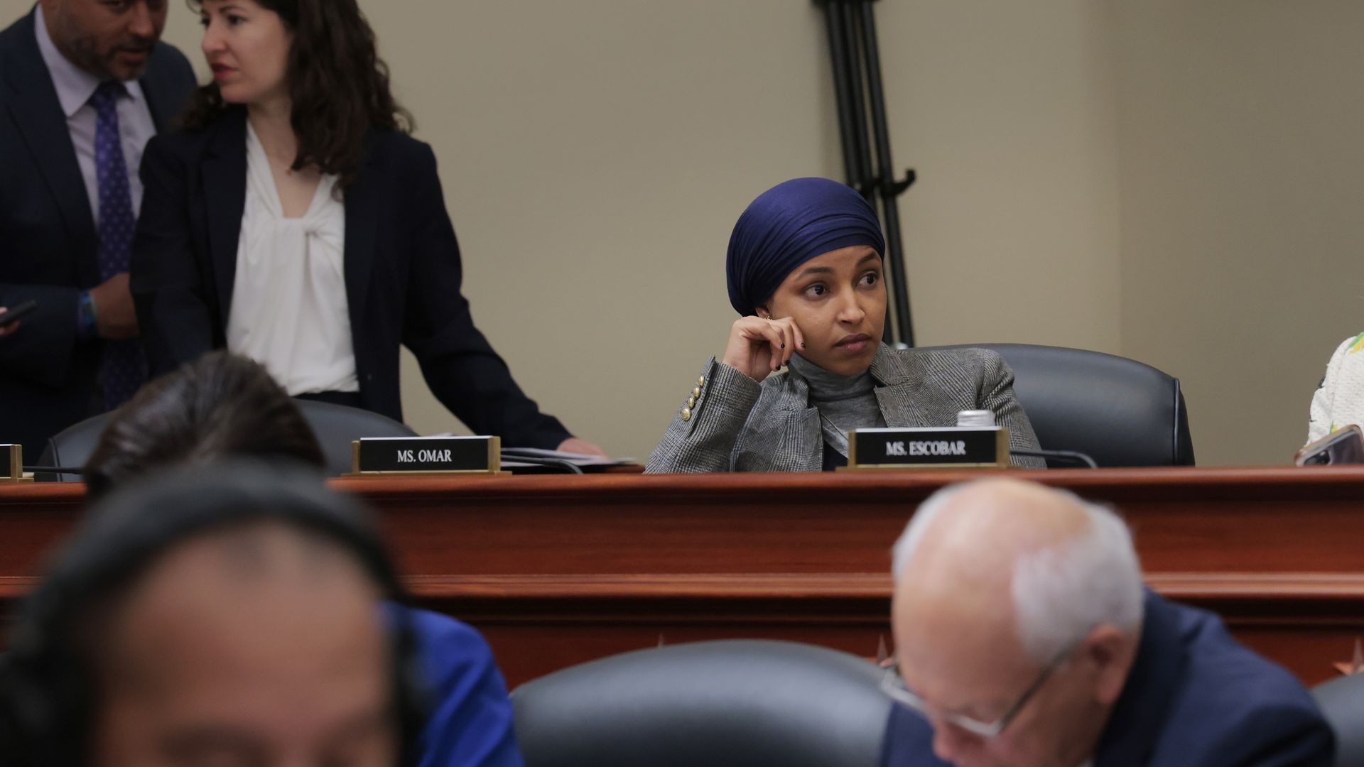 Rep. Ilhan Omar, wearing a gray suit and a blue headscarf, sits at a wooden dais with her face resting on her hand.