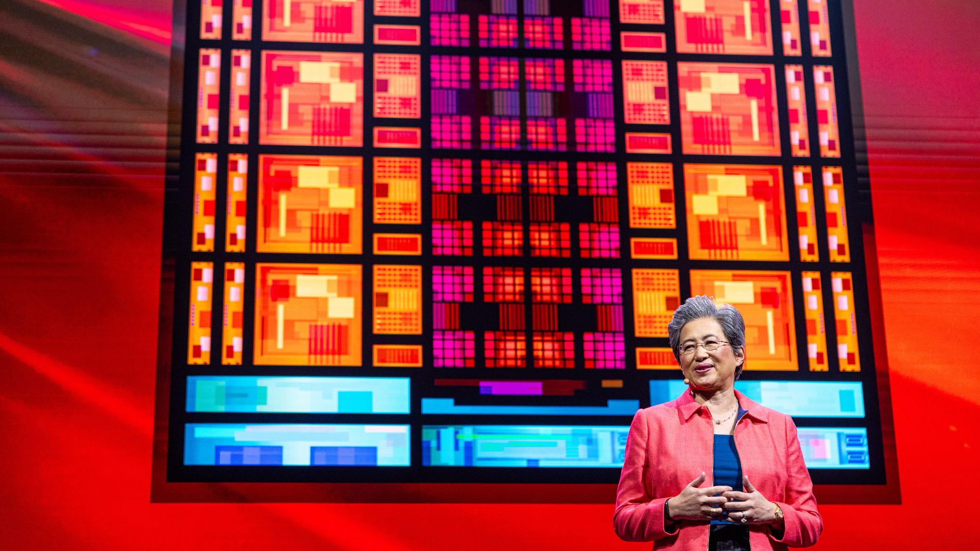 photo of a woman with short gray hair wearing glasses standing on stage against a screen showing a vibrant silicon chip