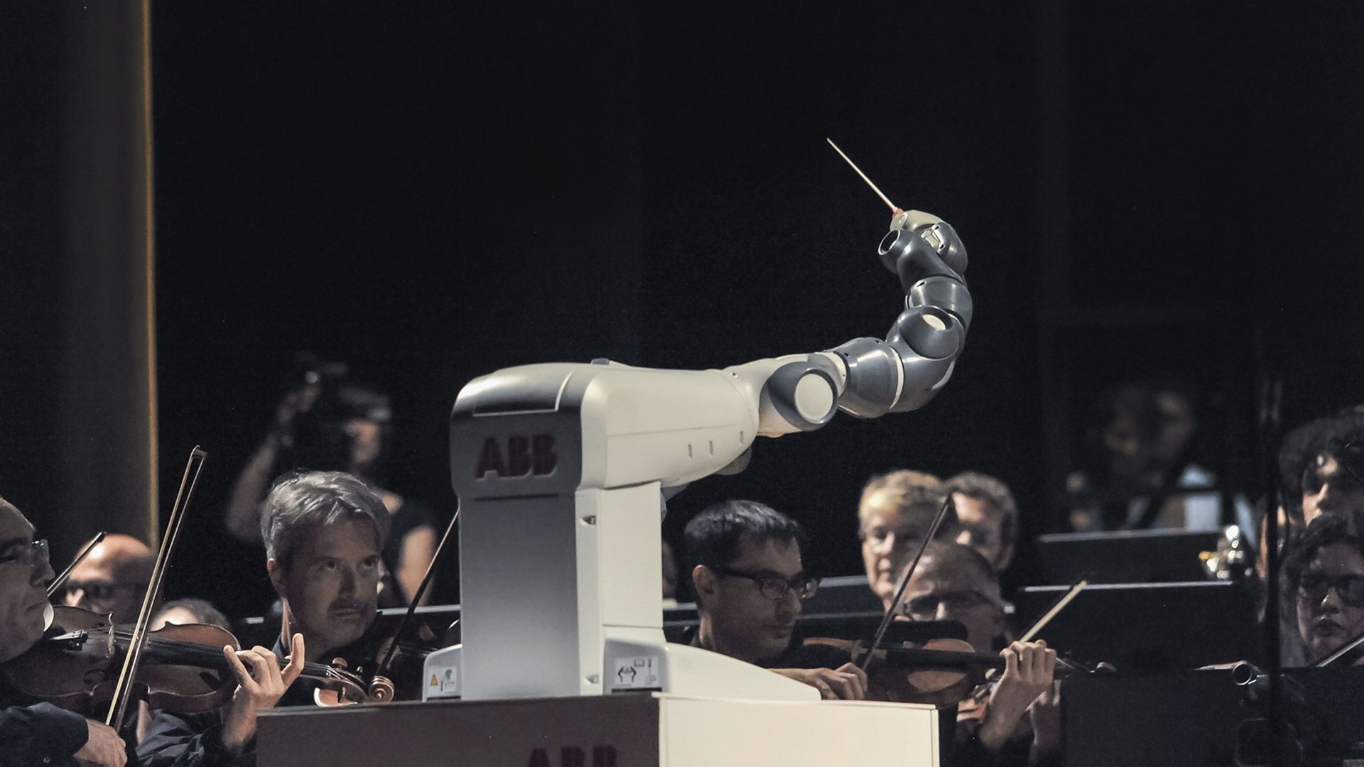 A modern photograph of a robot arm holding a conductors wand, directing a live orchestra. 