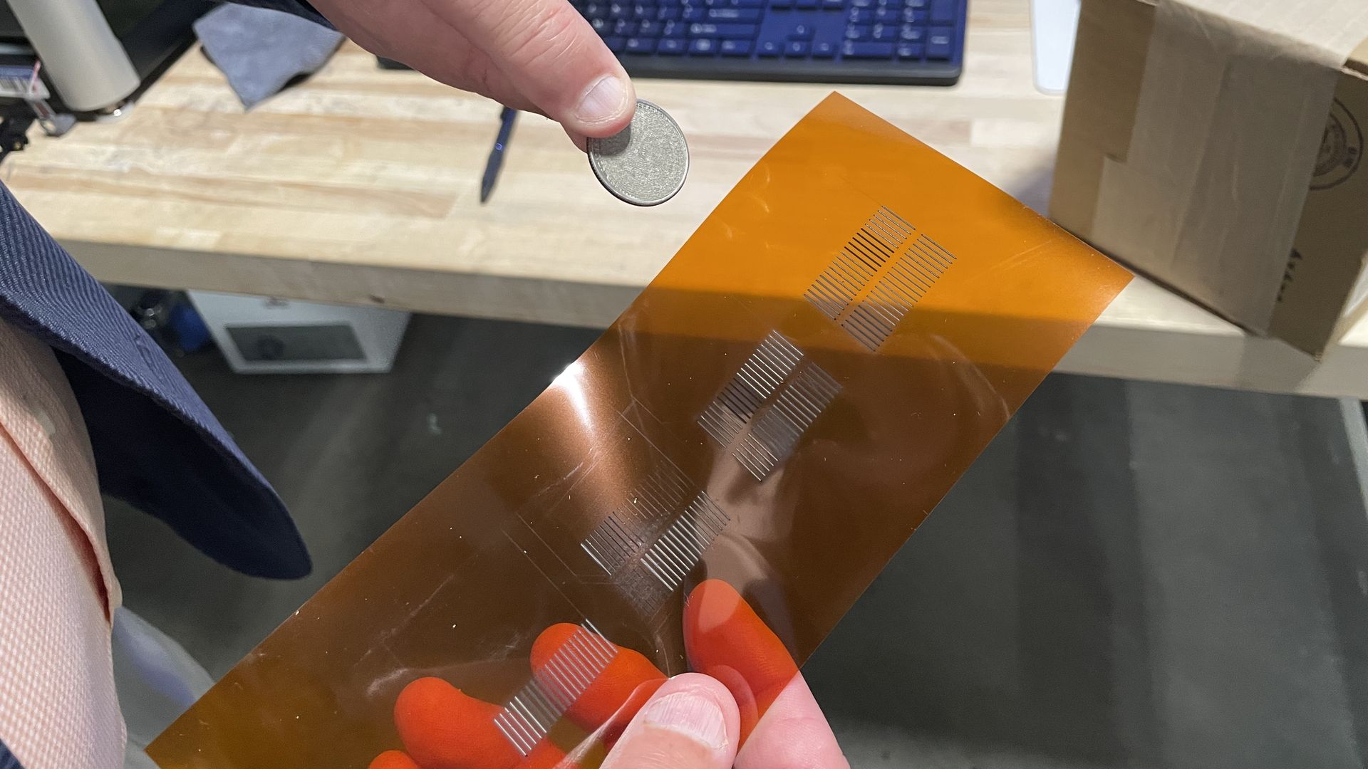 Close-up of a hand holding a small silver coin-shaped battery between the thumb and index finger over an orange flexible sheet on a wooden desk; a blue keyboard and box appear in the background.