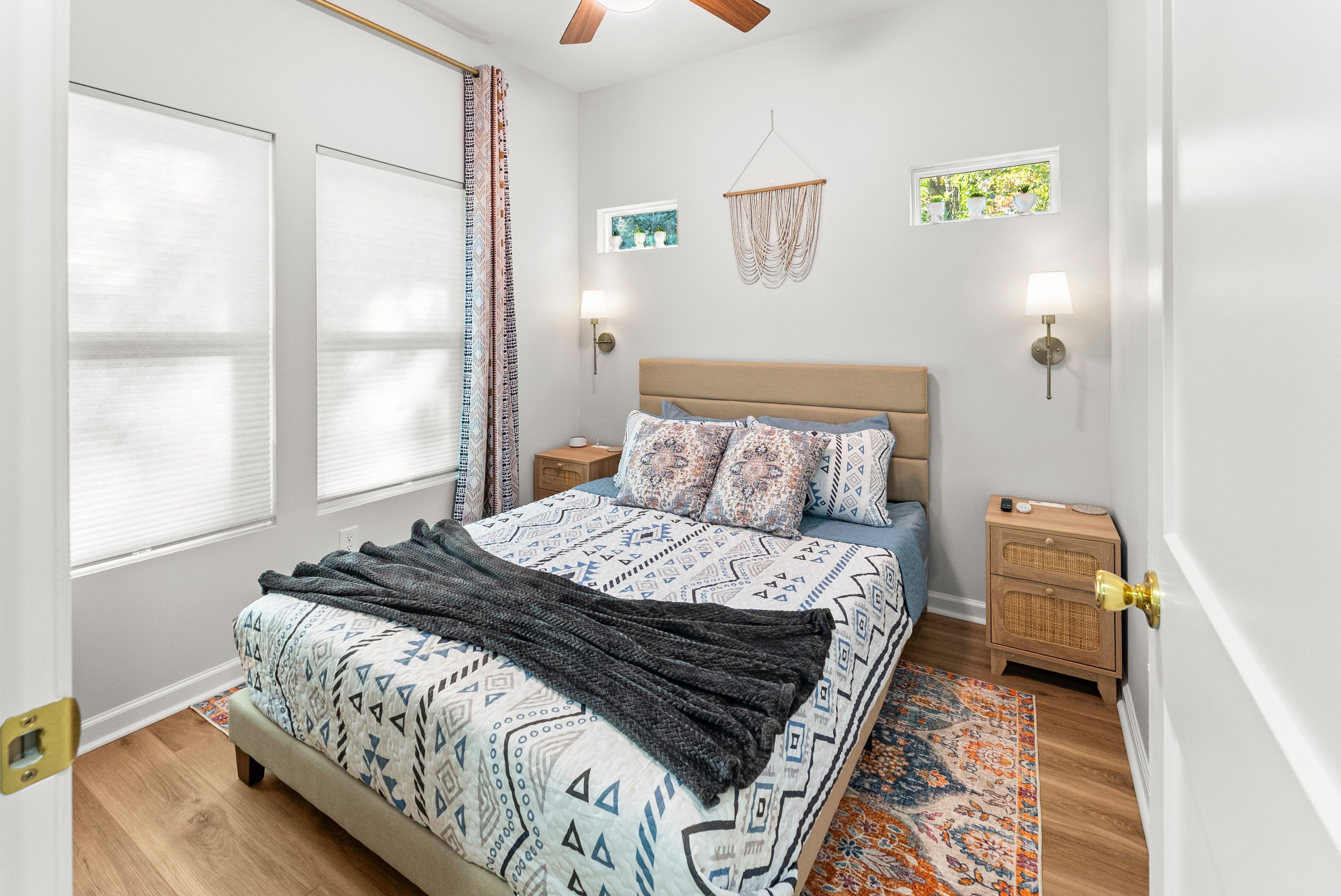 Bright bedroom with beige bedframe, patterned blue and white bedding, decorative pillows, dark gray blanket, wooden nightstands, wall lamps, and colorful rug on wood floor.