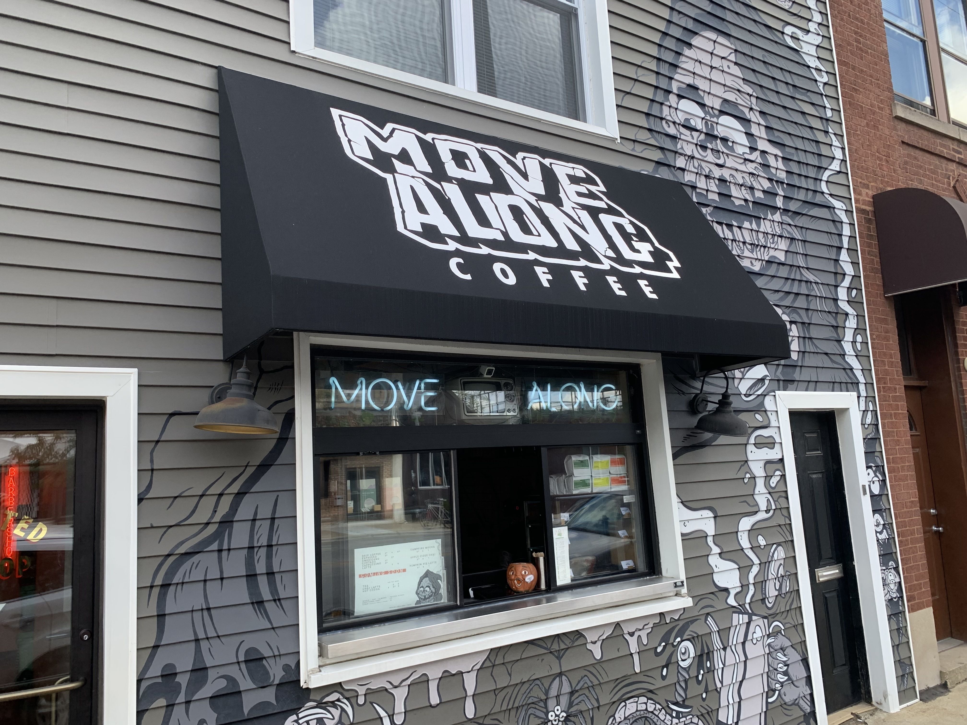 Black awning with white text "MOVE ALONG COFFEE" above a window with neon "MOVE ALONG" sign. Gray building has black and white mural featuring skull and abstract designs.