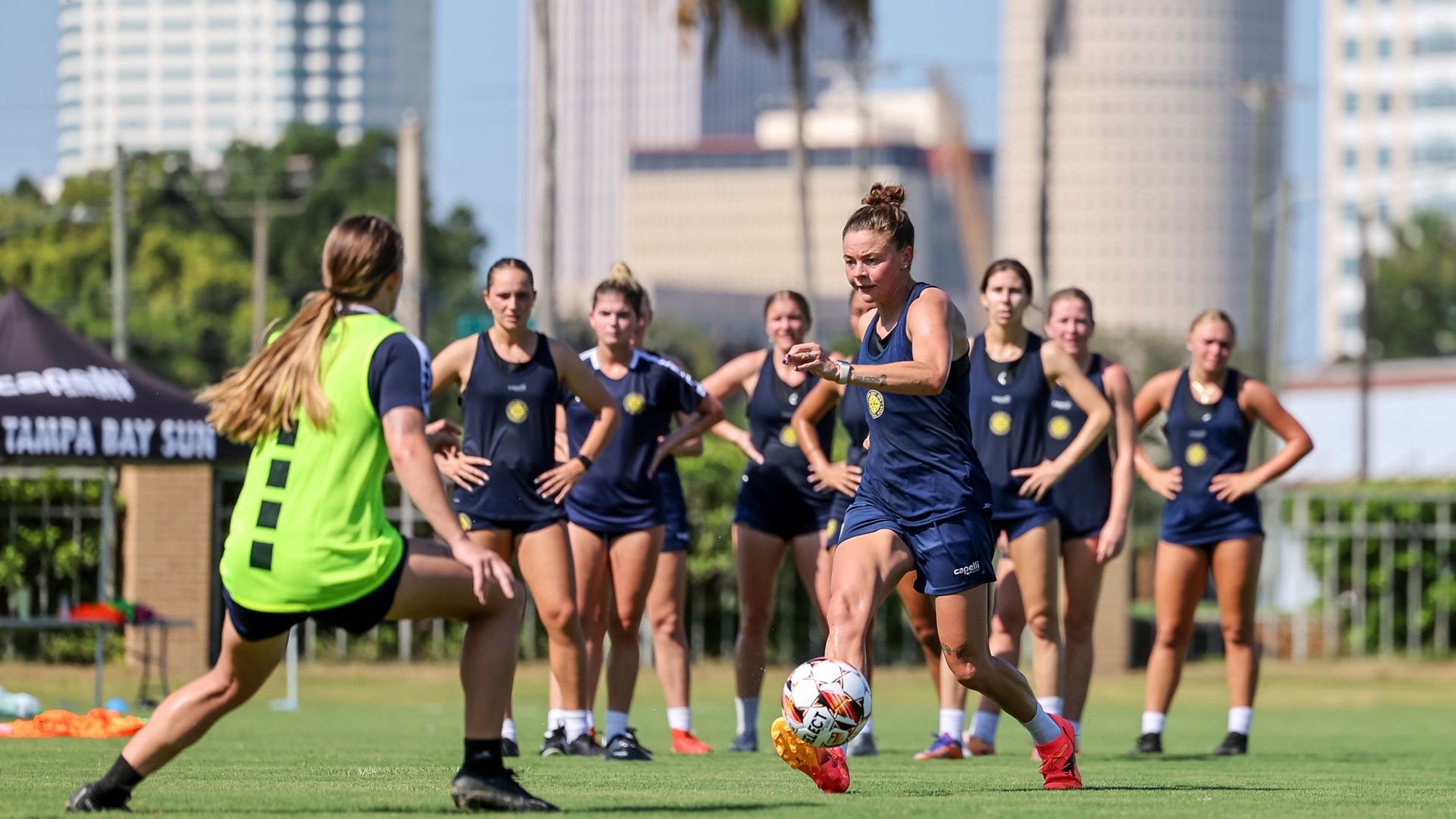 A soccer player dribbles a ball while her teammates, in navy jerseys, stand behind her. High-rise buildings that make up downtown Tampa rise in the background.