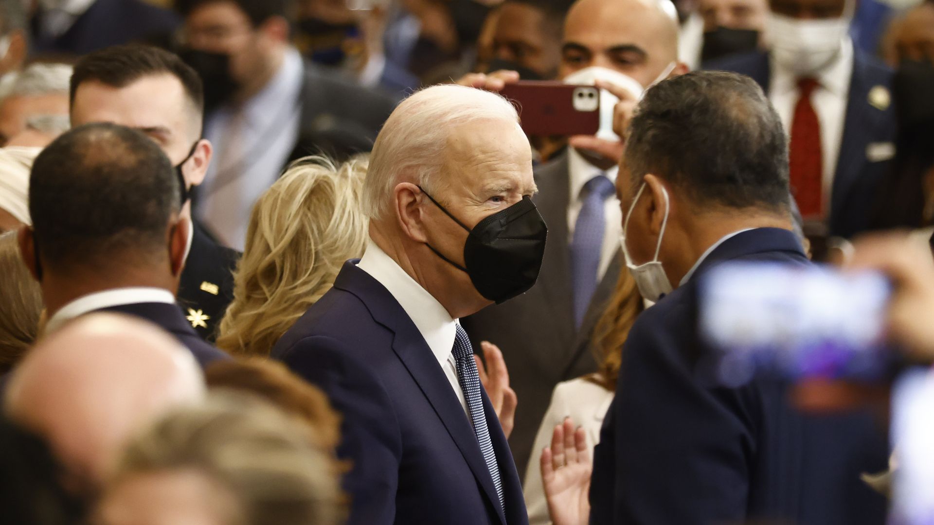 Photo of Joe Biden in a mask in a White House chamber surrounded by people