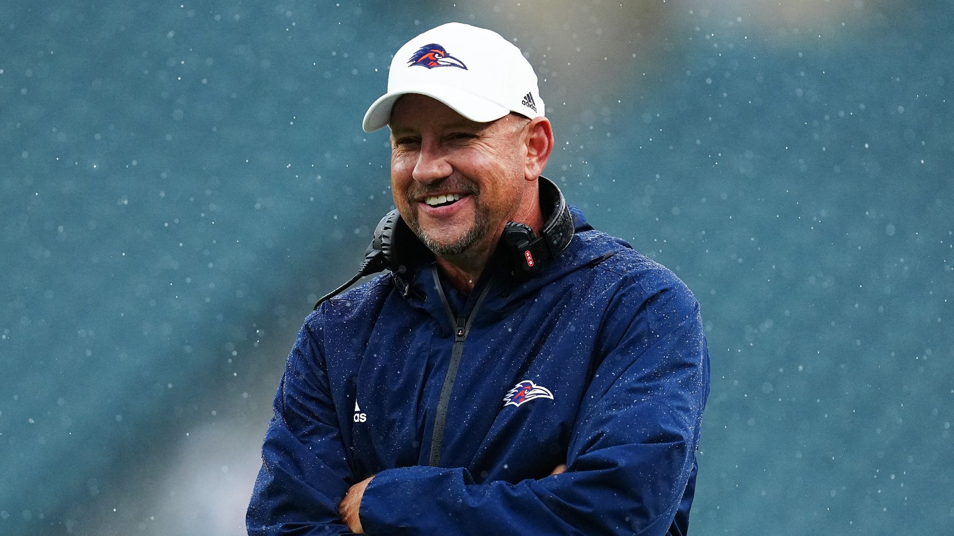 Jeff Traylor, dressed in a blue UTSA jacket and a white baseball cap, smiles at something away from the camera.