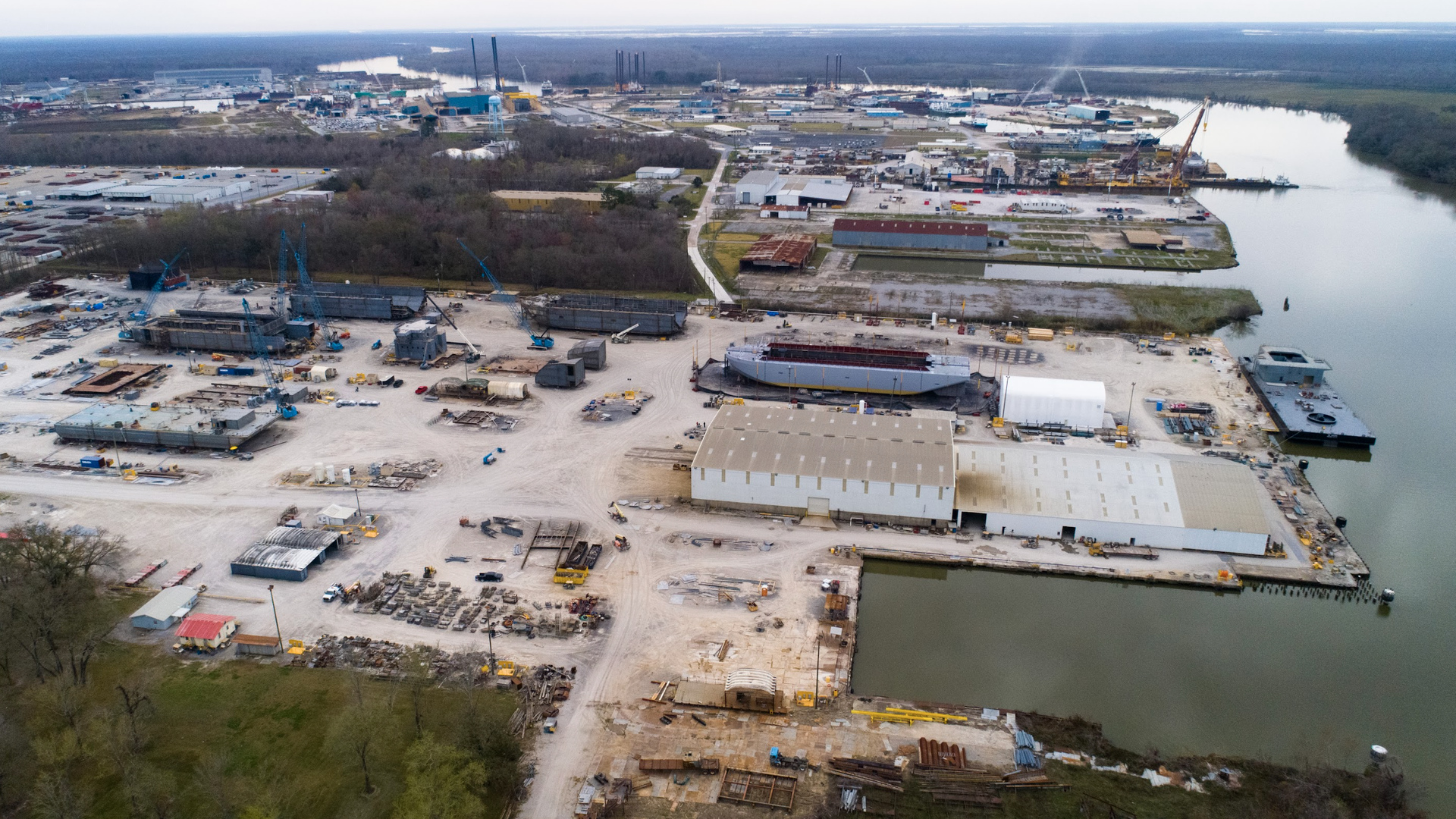 An aerial view of a Southern shipyard. Water is seen on the right side. Land and heavy machinery is on the left. The sky overhead is gloomy.