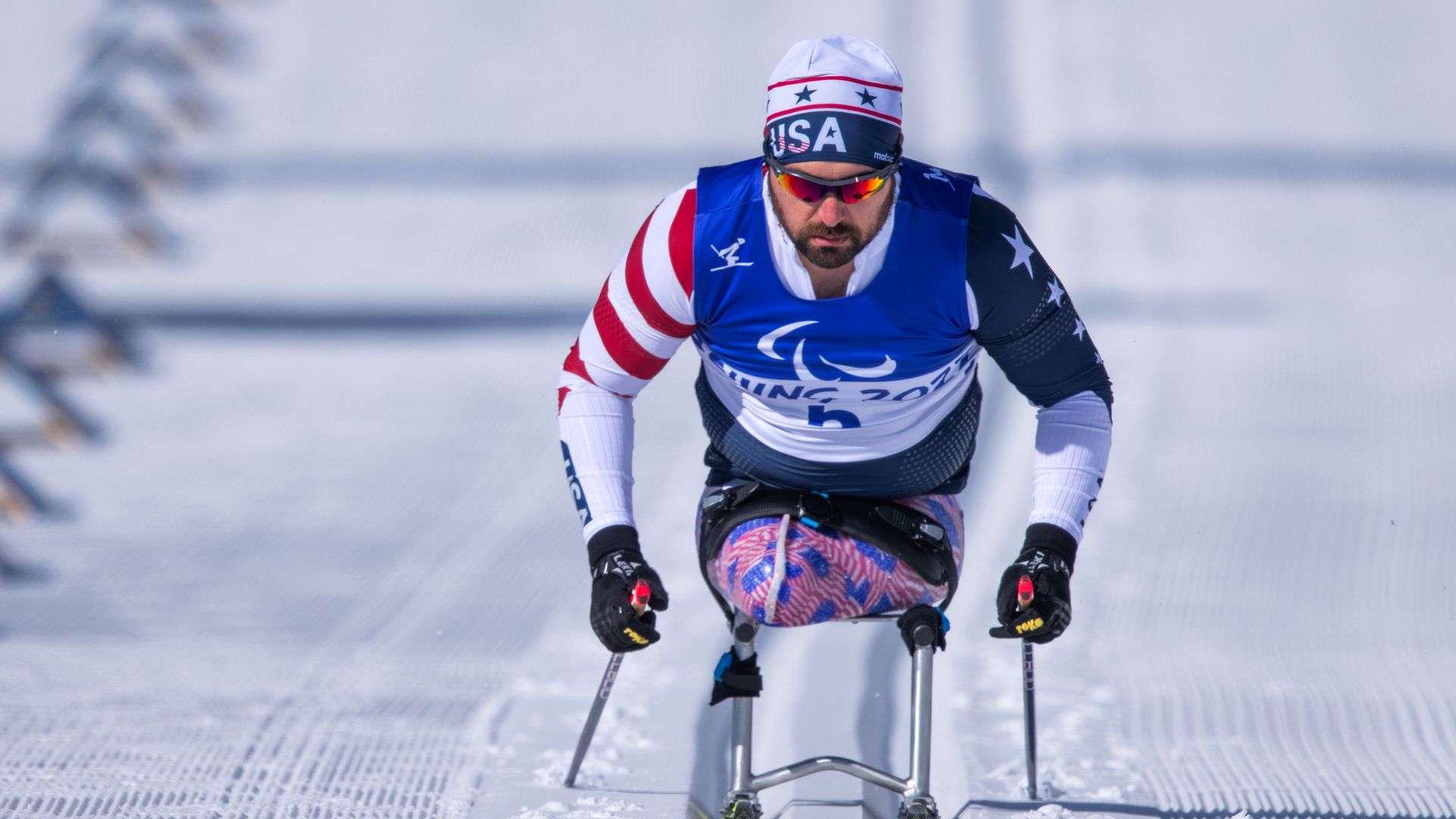 Paralympic skier from USA in blue bib skiing on snowy track using ski poles, wearing red, white, and blue outfit with stars and stripes.