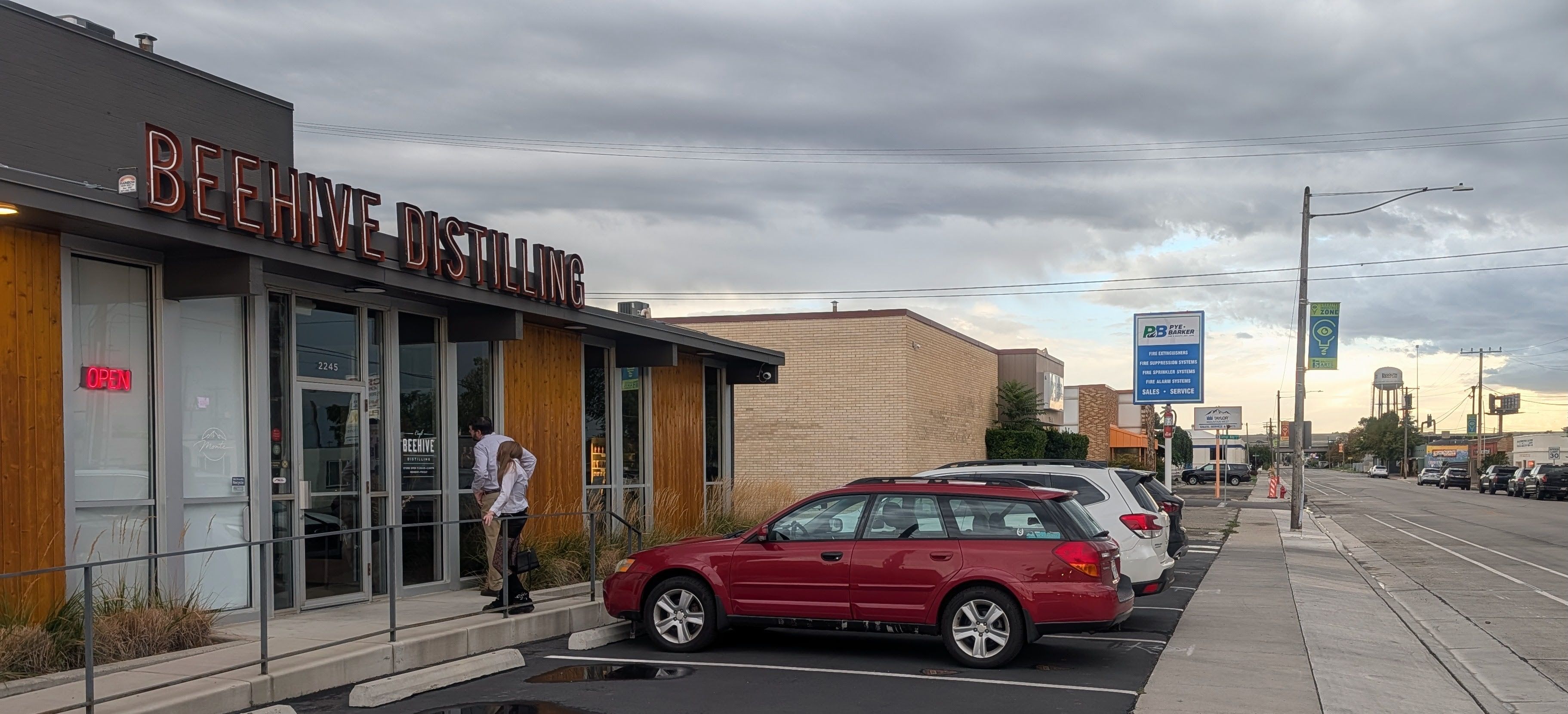 Exterior street view of Beehive Distilling with tall red sign on gray building, open neon sign, glass doors, two people entering, wooden panels, parked red SUV, overcast sky.