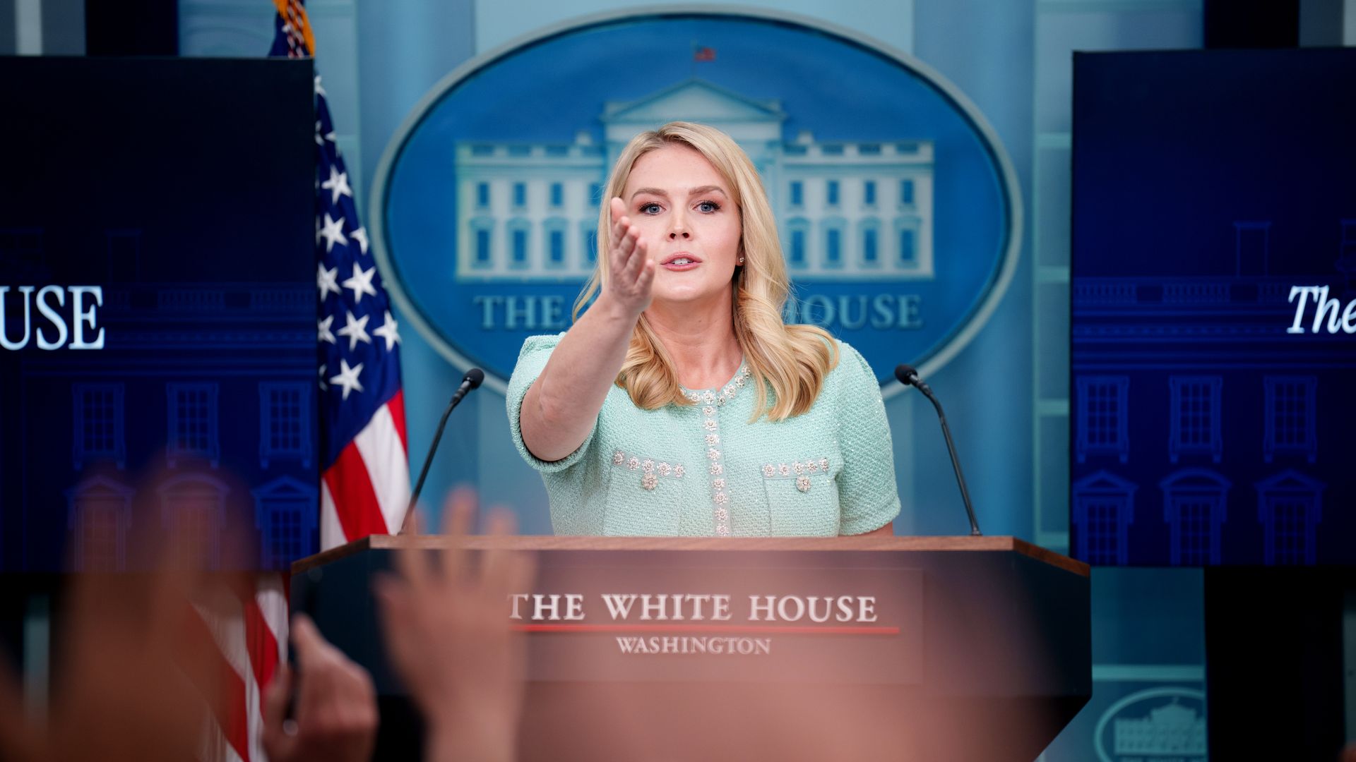 Karoline Leavitt stands at lectern with one arm outstretched. Several hands are seen raised in the foreground 