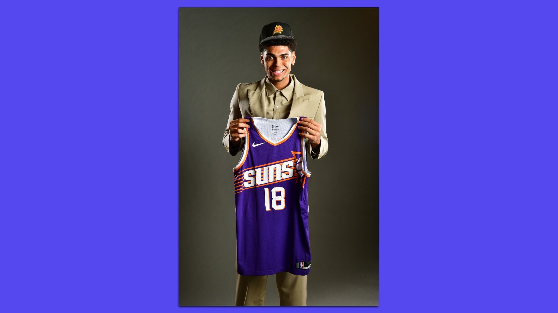 A young man in a tan suit and a black ballcap with a Phoenix Suns logo holding up a purple basketball jersey with "SUNS" and the number 18 on the front; he's standing in front of a dark gray background. 