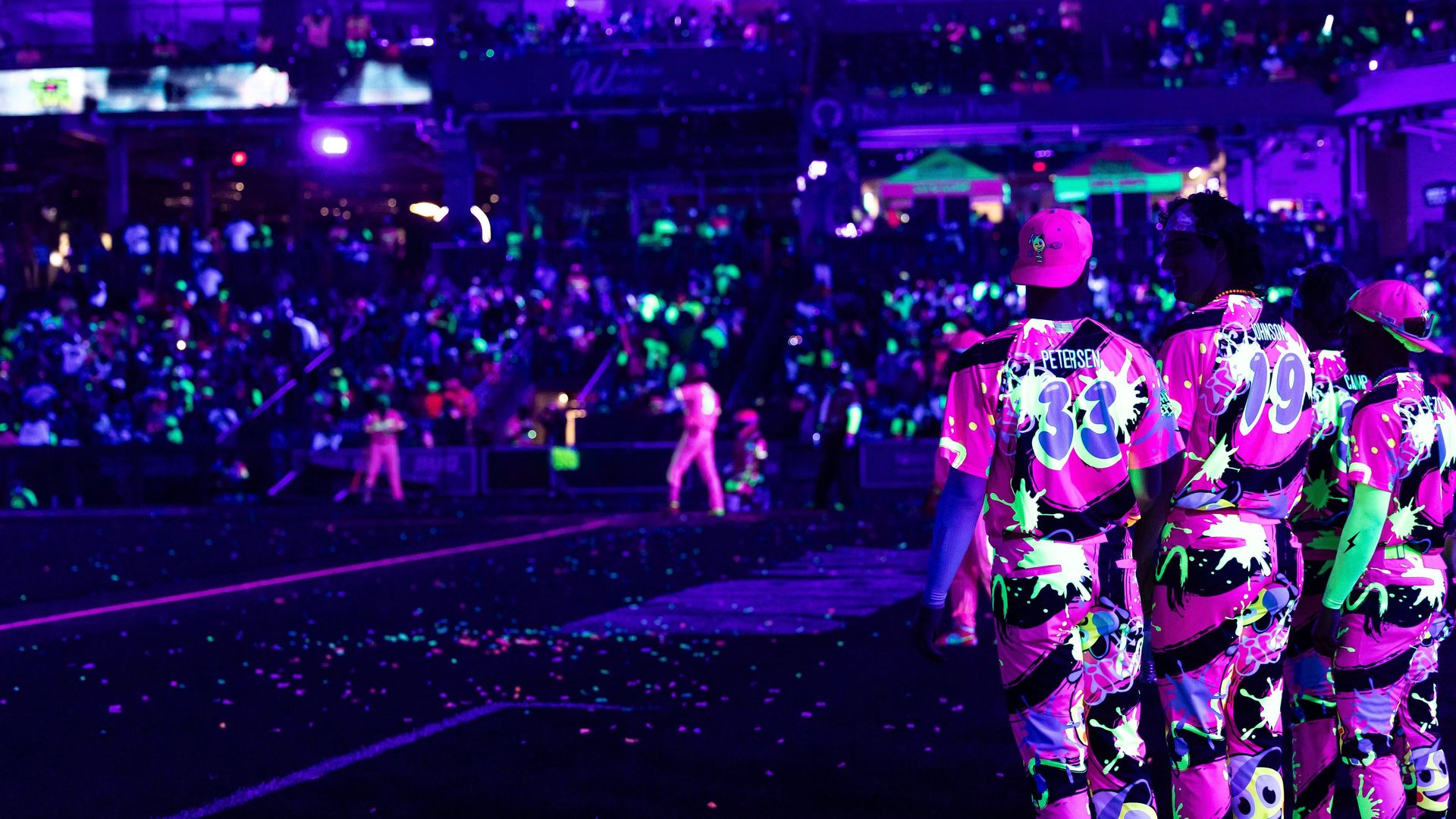 Players in neon pink and black baseball uniforms with glowing paint effects stand in a dark stadium under purple lights, with a crowd and players visible in the background.