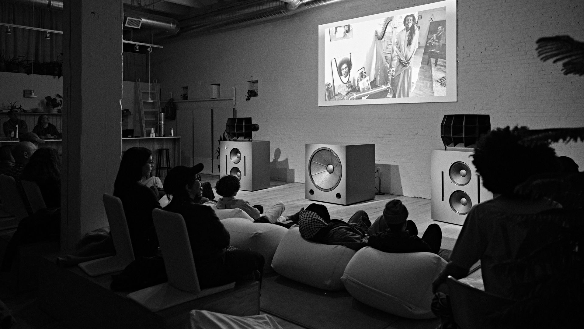 Dim industrial room with exposed ducts and large speakers; audience sits on beanbags and chairs, watching a projected black-and-white photo on the wall.