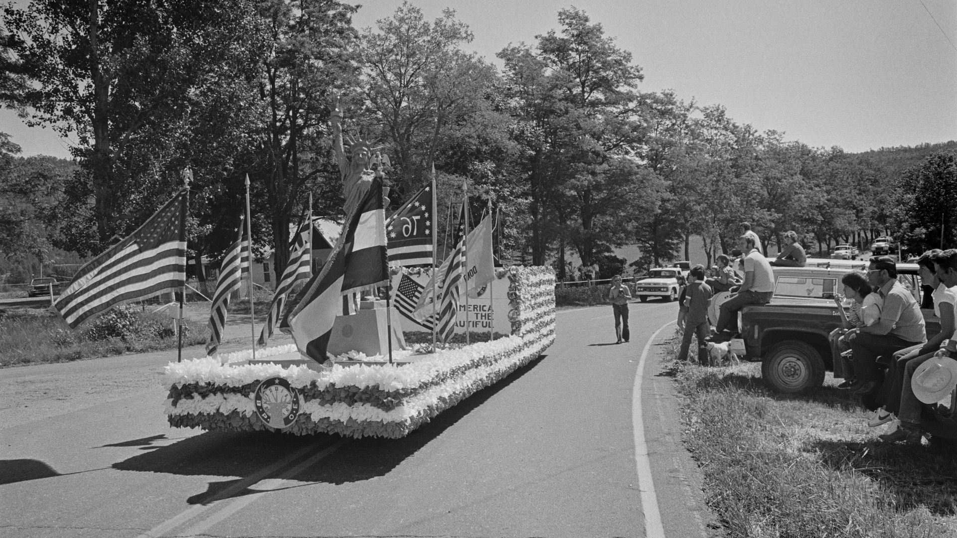 A parade float with American flags travels down a road as people off to side watch on, sitting on the backs of cars and trucks.