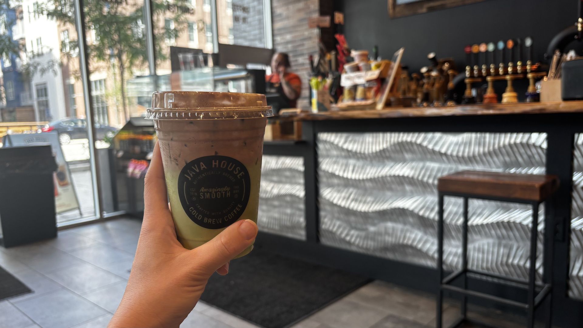 Hand holding a cold brew coffee cup with a black lid inside a coffee shop with wavy silver panels, wooden counter, bar stool, and a blurred barista in the background.
