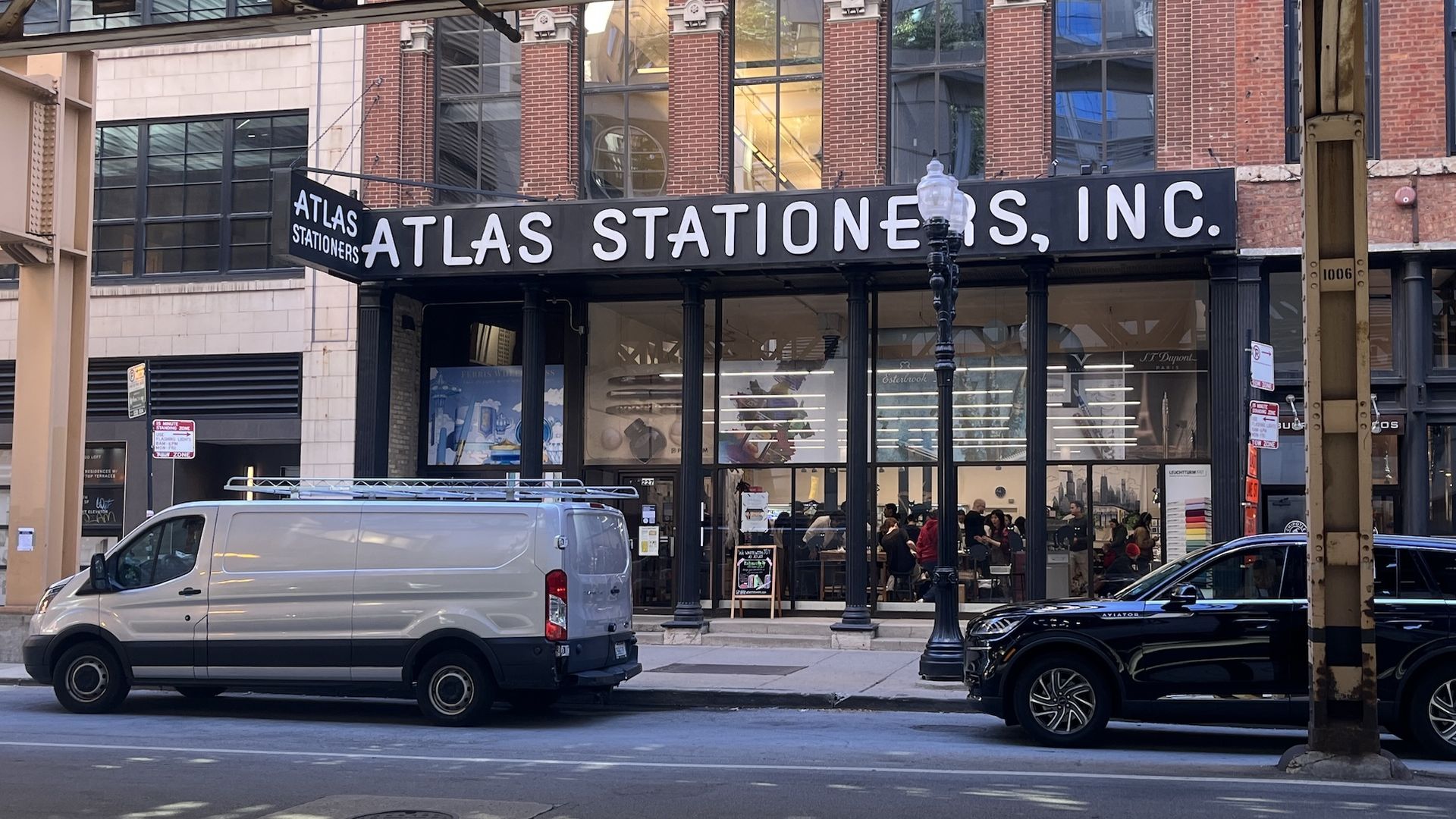 Exterior of Atlas Stationers, Inc. store with large black and white sign on a brick building, two parked vehicles in front, and people visible inside through large windows.