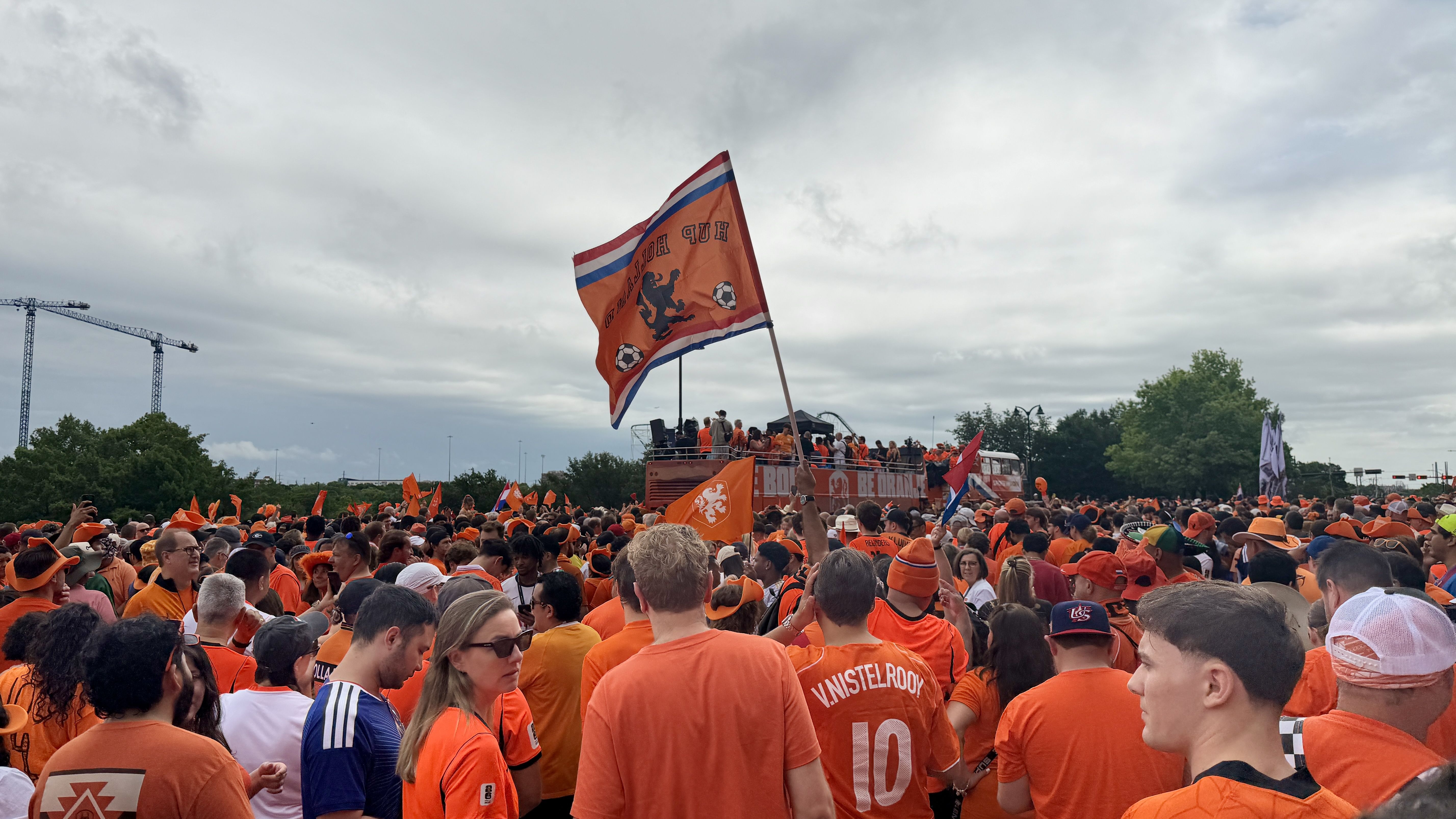 Massive crowd of orange-clad fans at a rally, waving a large orange flag with a lion and soccer balls on a float. Overcast sky, construction cranes in the background.
