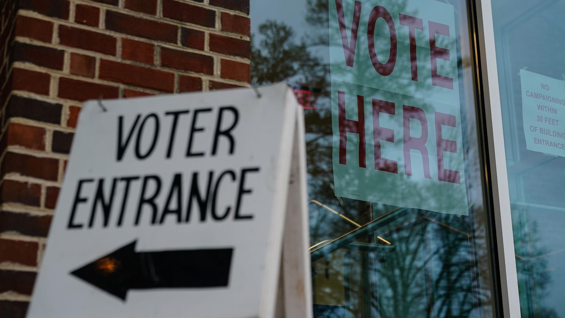 A sign propped on the floor reads "VOTER ENTRANCE" with an arrow pointing left. A poster on a window behind reads "VOTE HERE."
