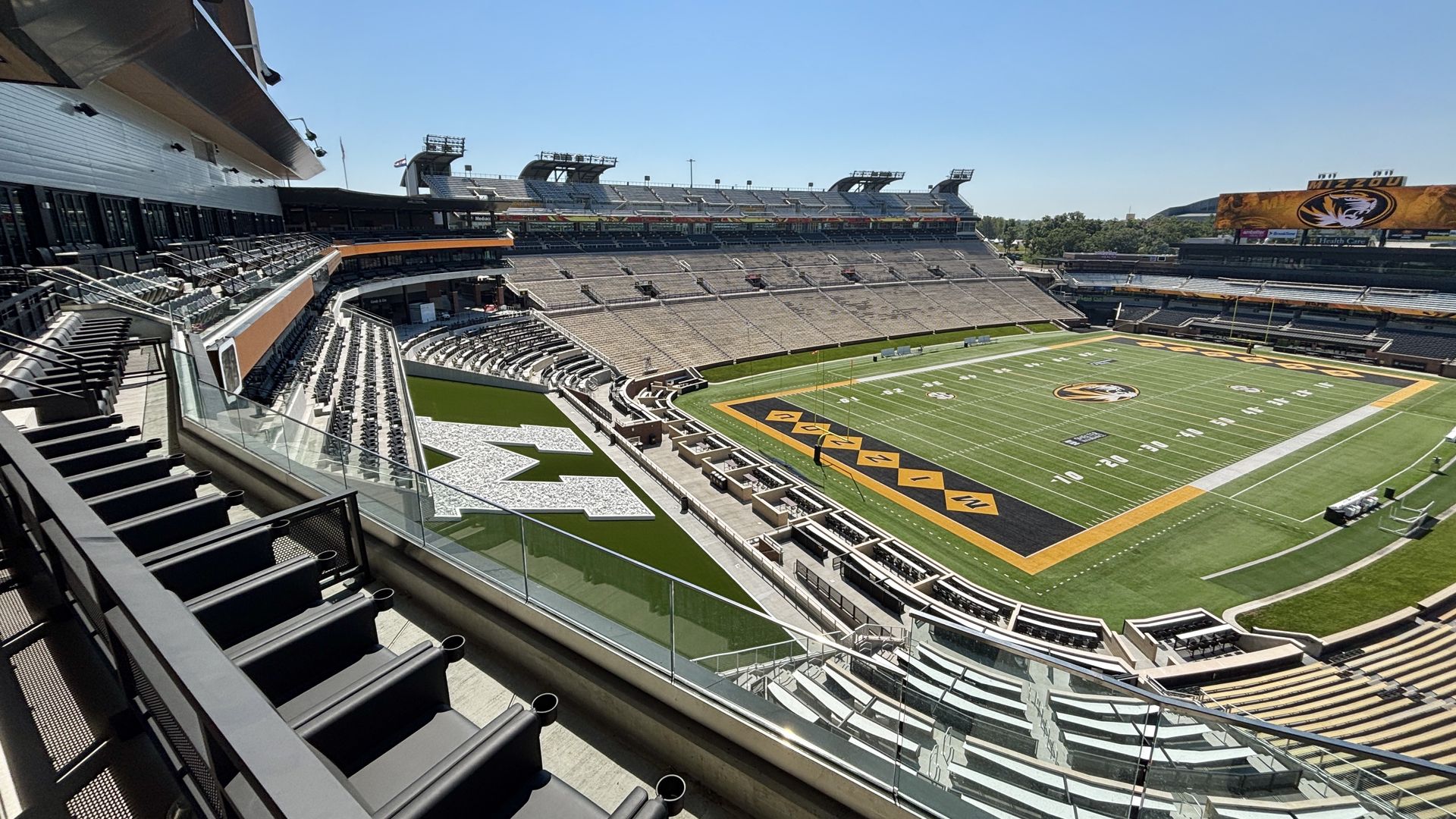 Wide view of an empty college football stadium with green field, yellow end zones, and the MIZZOU tiger logo. Grey bleachers, glass railings, under a clear blue sky.