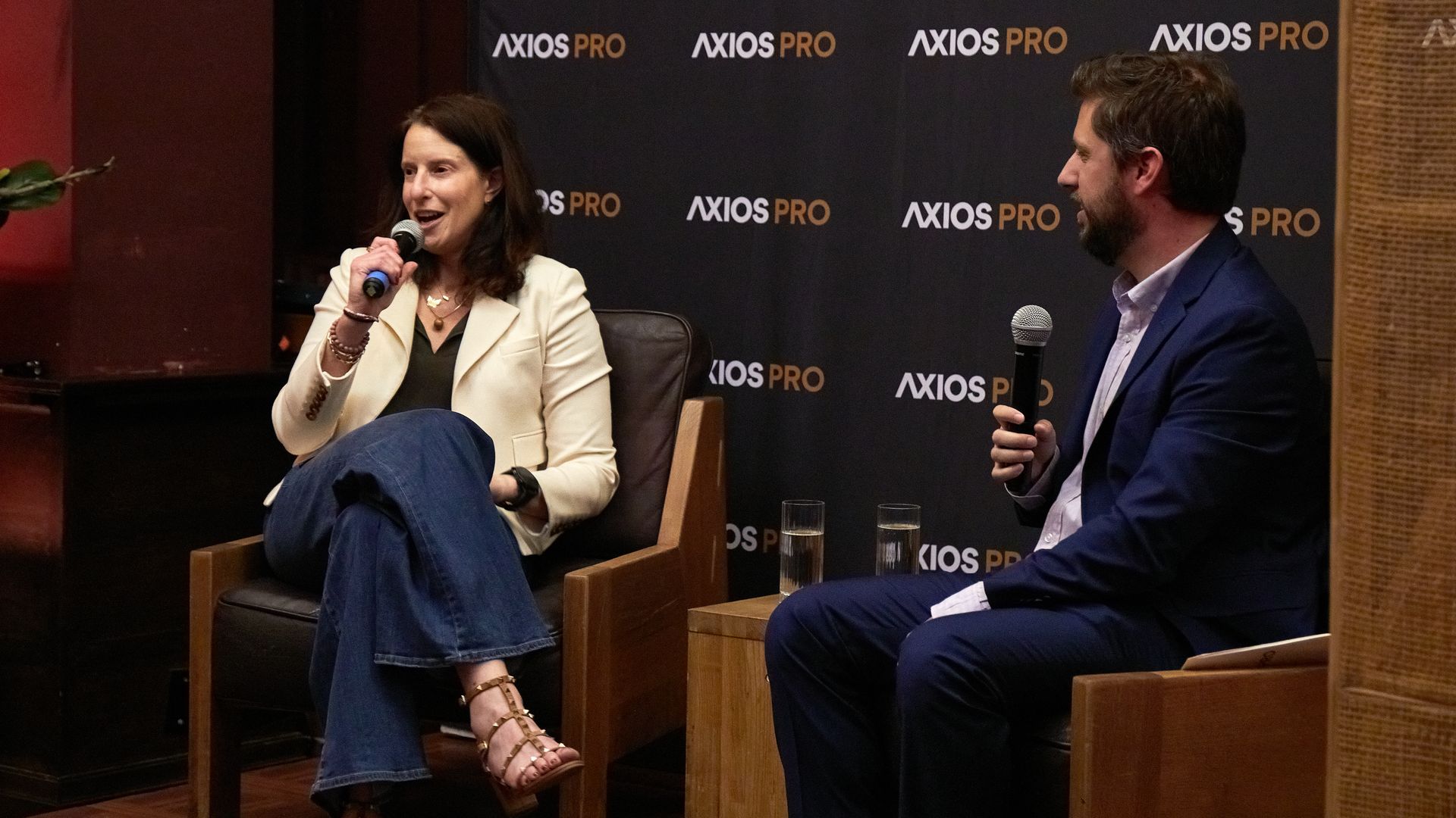Kara Nortman in a cream blazer and jeans and Tim Baysinger in a blue suit sitting on chairs, holding microphones in front of a backdrop that says Axios Pro