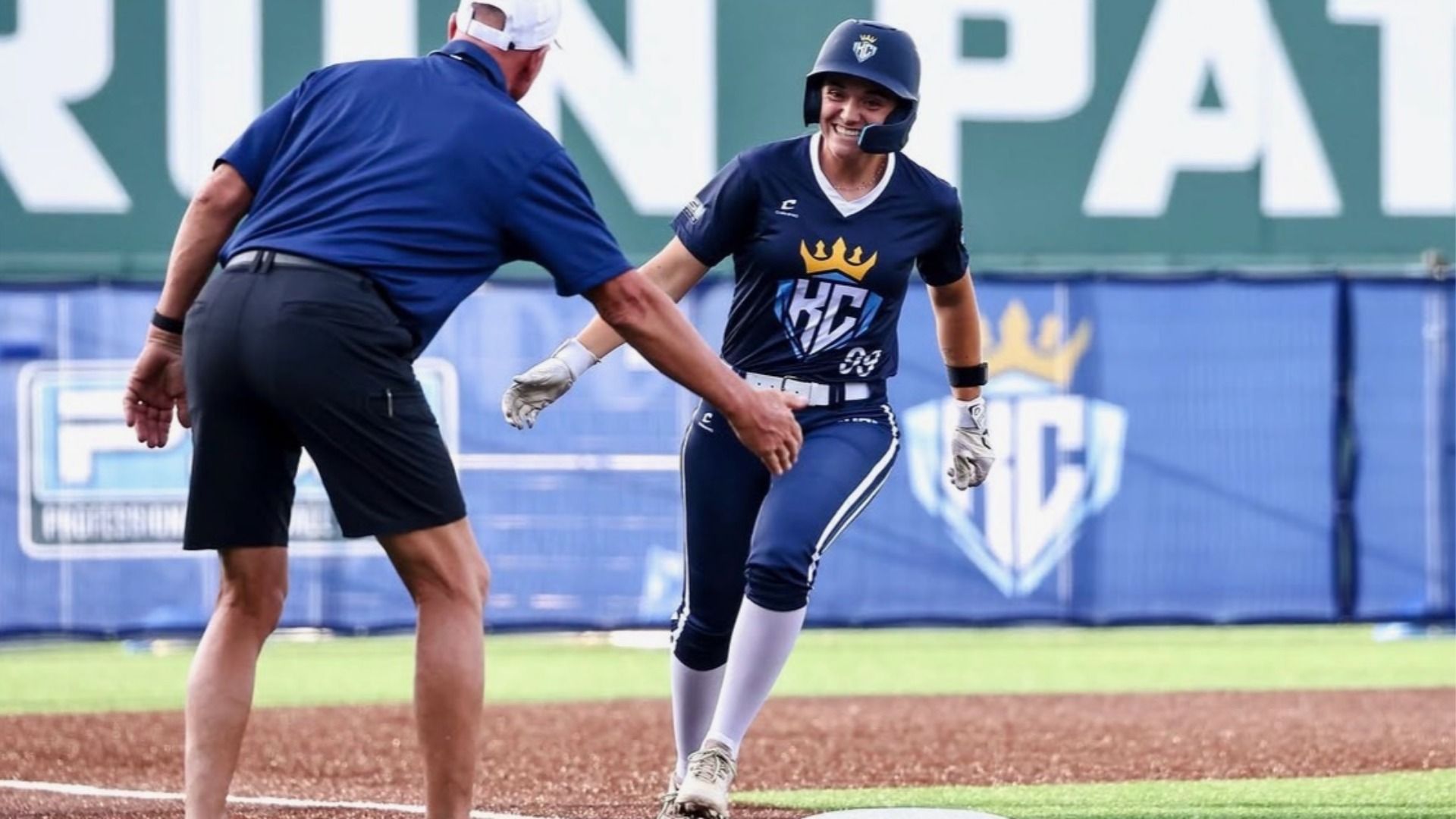 Smiling female softball player in navy uniform with the KC Diamonds logo, including a crown and a "KC", also wearing a helmet, high-fiving a base coach on a dirt infield; blue outfield banners and large white letters visible in the background.
