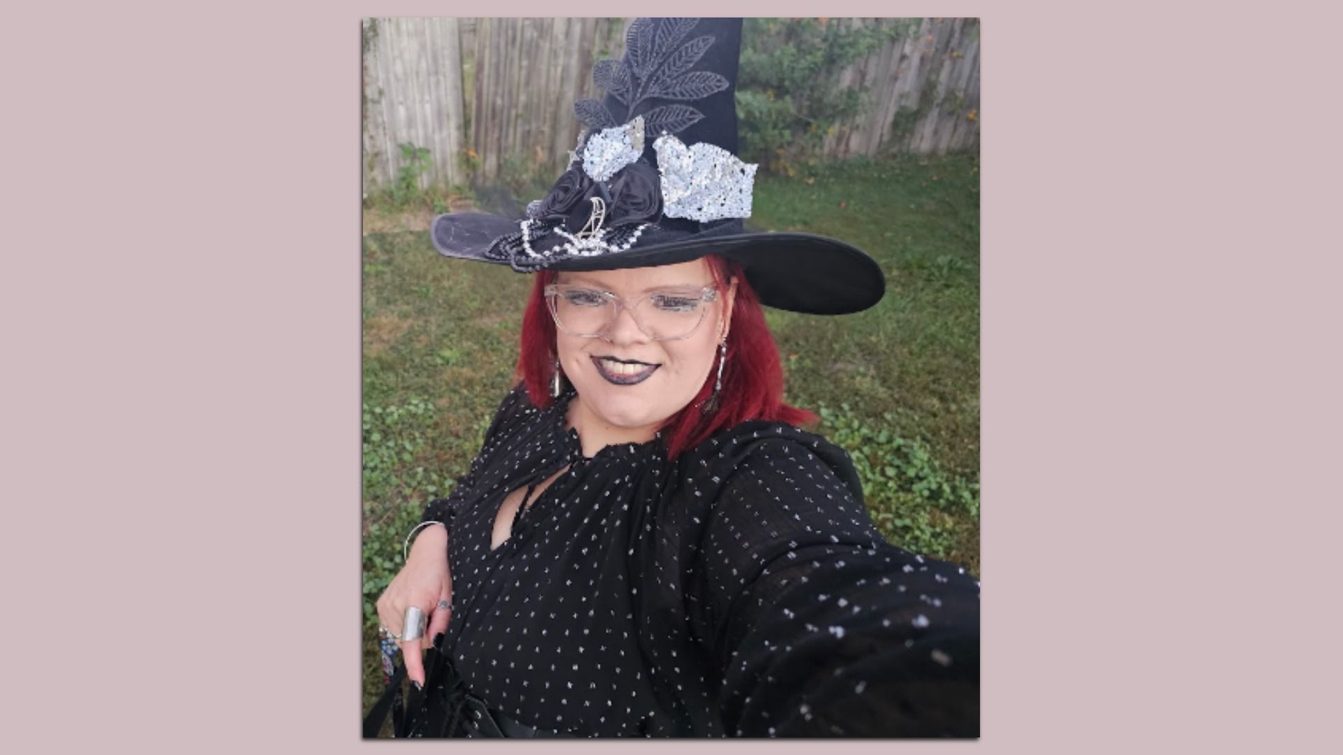 Person with red hair wearing a tall black witch hat adorned with black and silver decorations, black lipstick, clear glasses, and a black dress with white dots, outdoors on grass.
