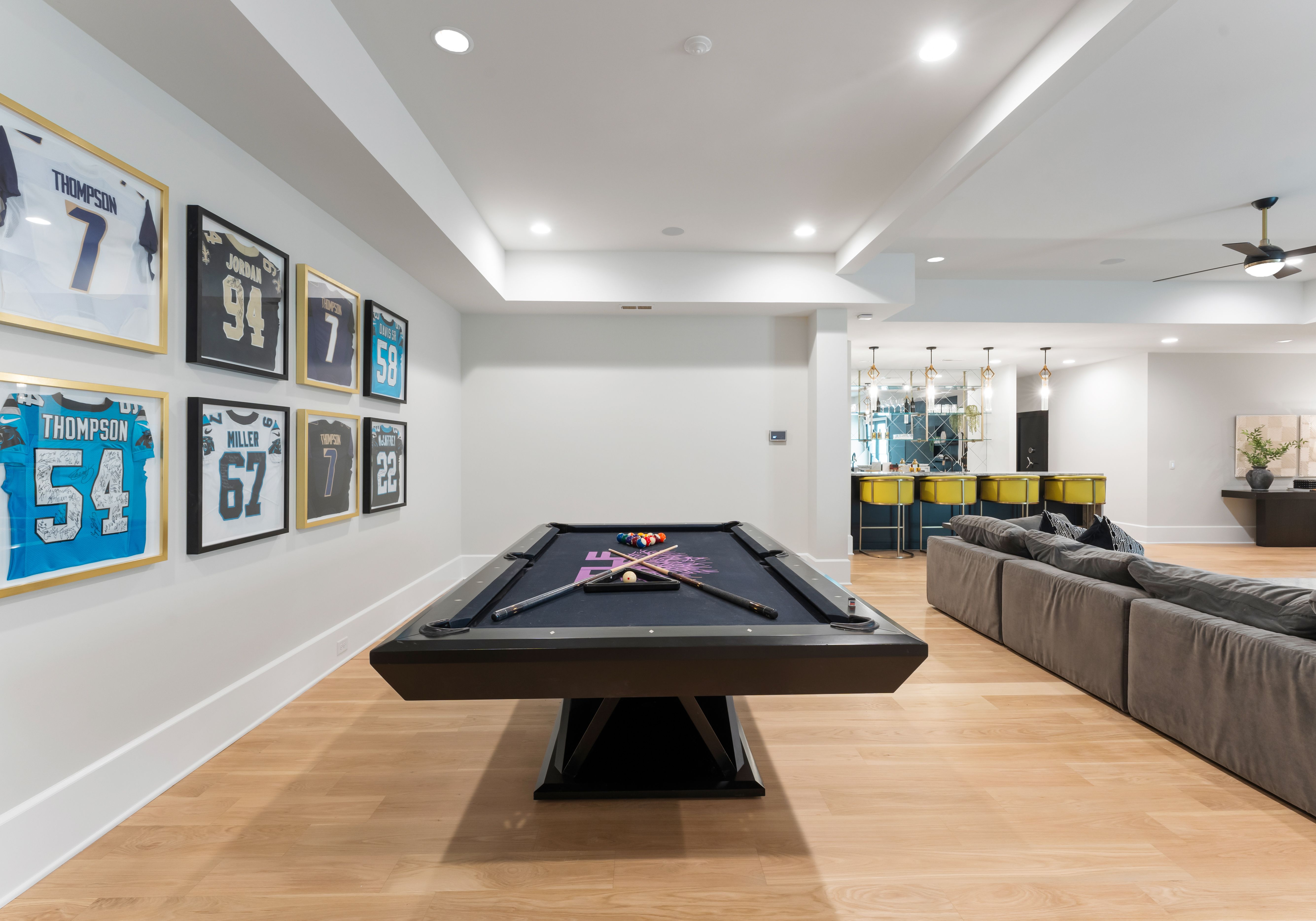 Modern basement game room with a black pool table, framed sports jerseys on white wall, gray sectional sofa, wooden floor, and a bar with bright yellow stools in the background.